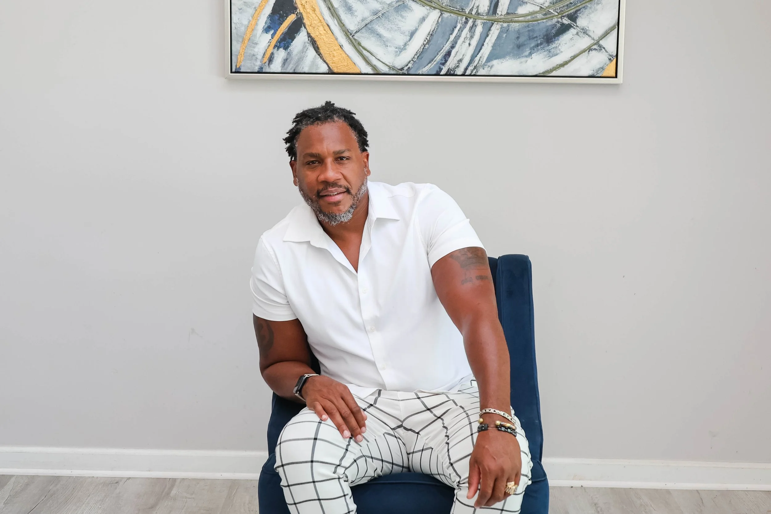 A middle-aged man with dark skin, gray beard and dreadlocks, sitting on a blue chair in a room with light gray walls and a framed abstract painting behind him. He is wearing a white short-sleeved shirt and white plaid pants, with bracelets and rings on his left hand.