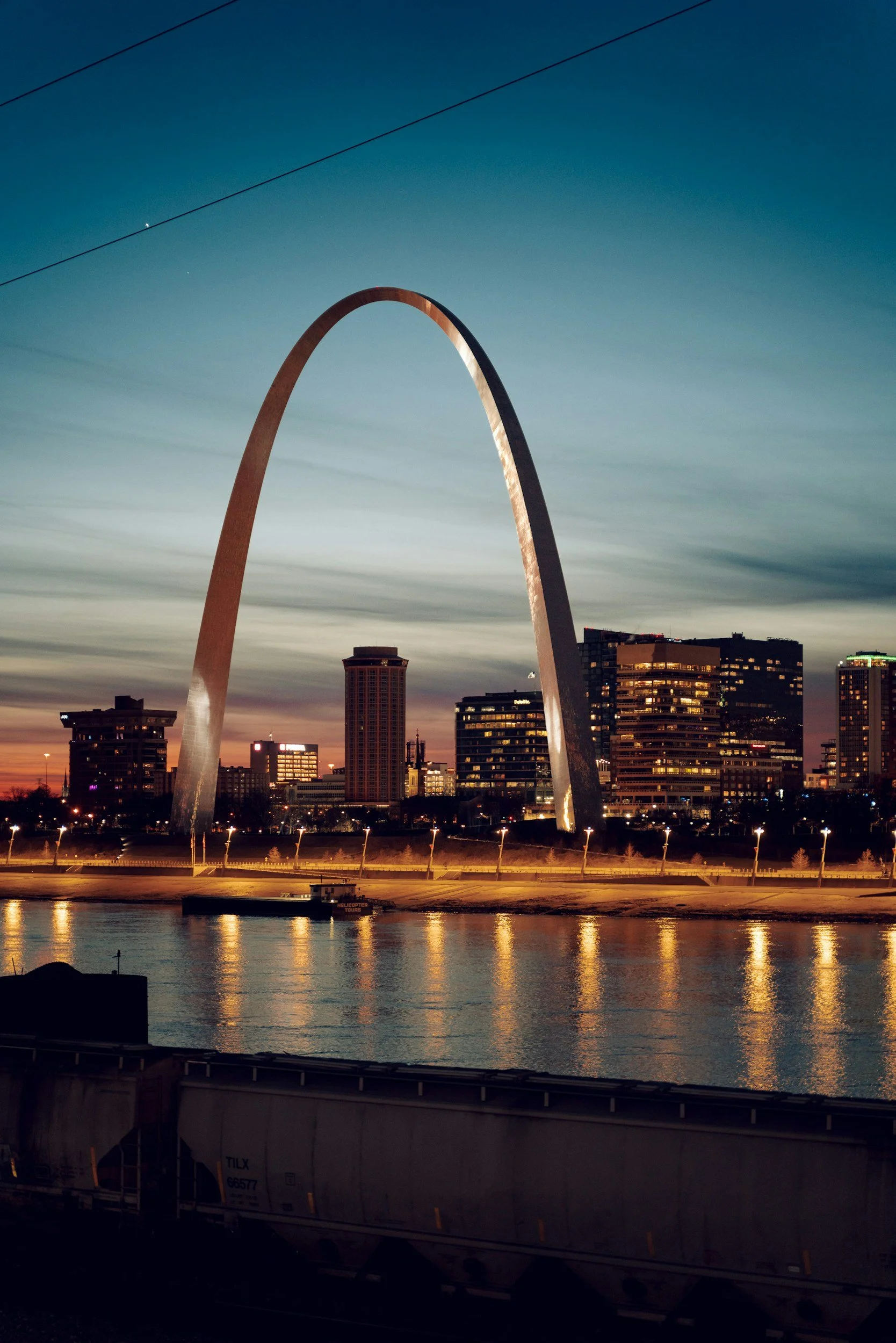Nighttime view of the Gateway Arch in St. Louis, Missouri, with city skyscrapers in the background and the river in the foreground reflecting lights.
