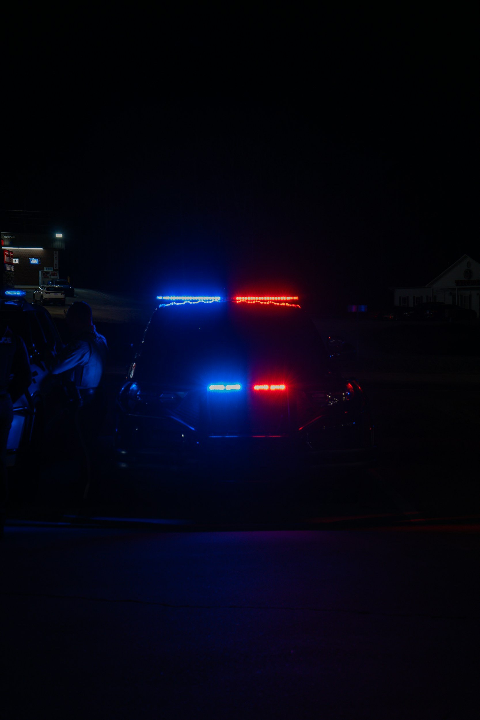 Police car at night with flashing blue and red lights, several people near the vehicle, and a building with signs in the background.