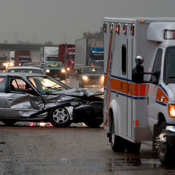 A car accident scene with a heavily damaged silver car and an emergency ambulance on a wet road, with traffic in the background.