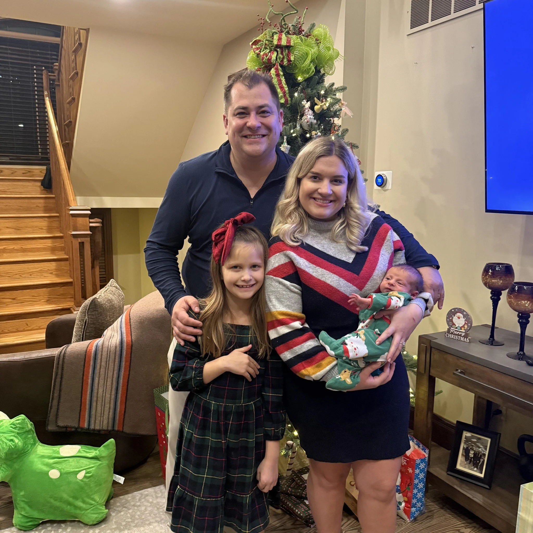 A family of four posing in a living room decorated for Christmas, with a Christmas tree and presents around them. The group includes a man, a woman holding a baby, and a young girl standing in front. They are smiling and dressed festively.