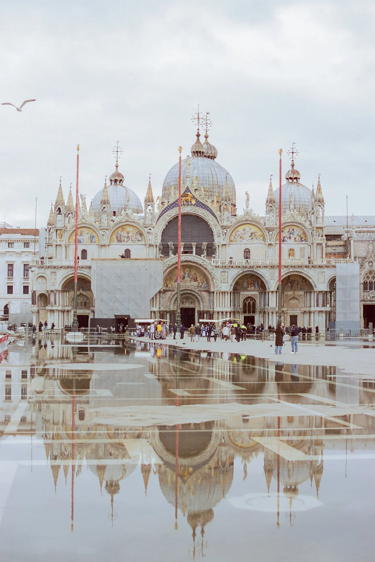 A historic cathedral with domes and ornate architecture, reflected in a large puddle in foreground, with cloudy sky above and people walking nearby.