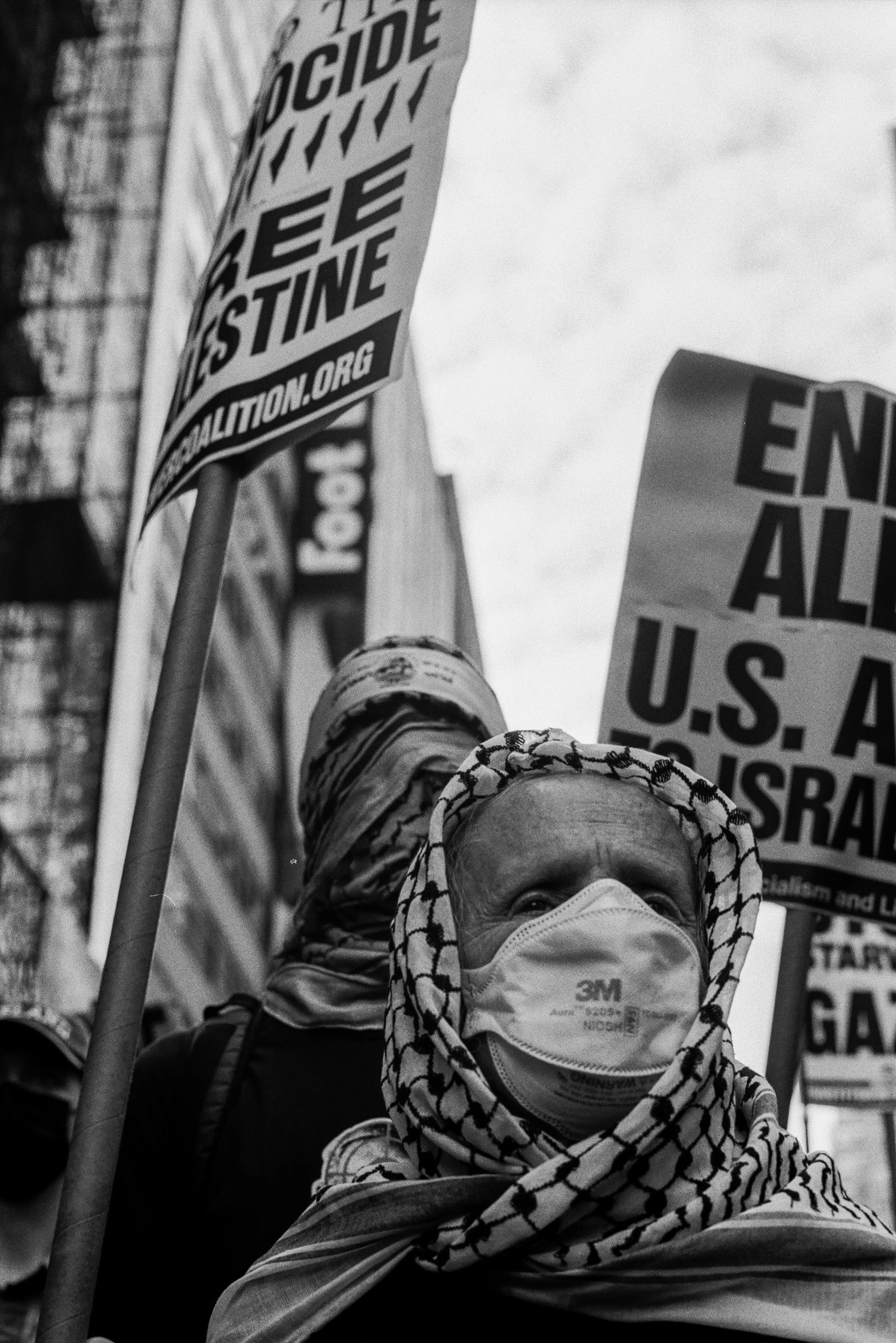 A person wearing a patterned headscarf and a face mask holding up protest signs in an urban setting. The signs include messages about justice, ending U.S. interference, and other political statements. The image is in black and white and appears to have been taken during a demonstration or protest.
