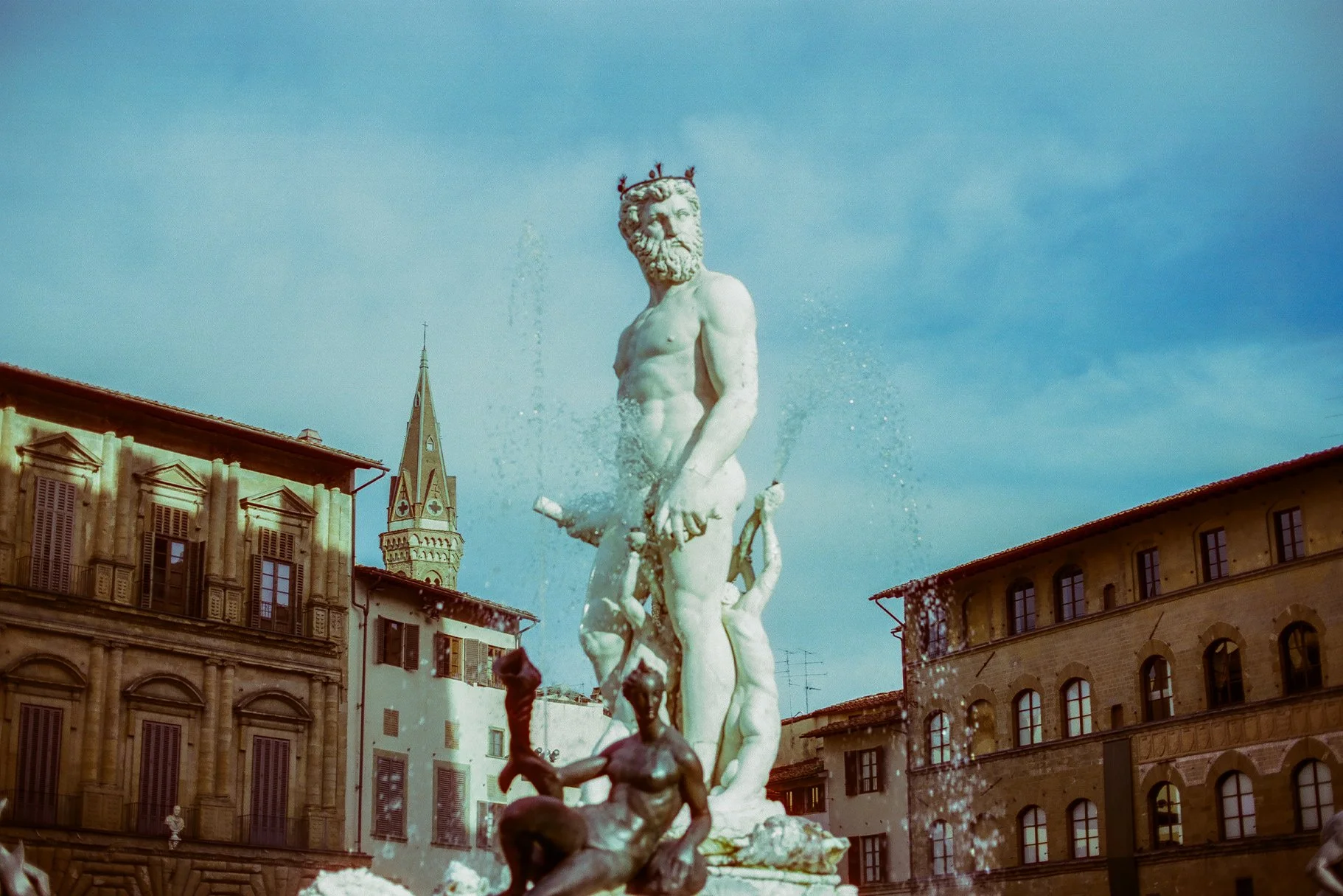 Fountain with a large marble statue of a bearded man with long hair, water spraying from the fountain, and surrounding historic European buildings with multiple windows.