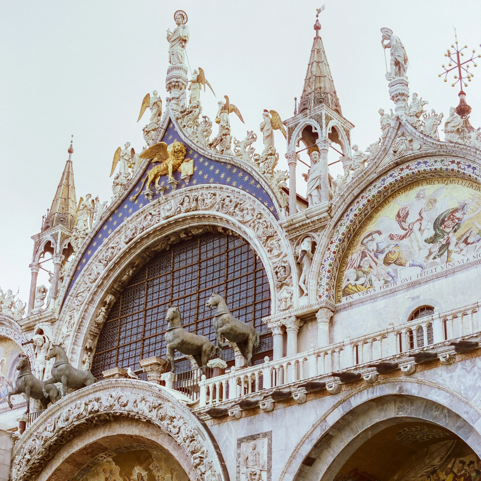 Close-up of the ornate façade of St. Mark's Basilica in Venice, Italy, featuring sculptures of horses, angels, and religious figures, with a mosaic above the archway.