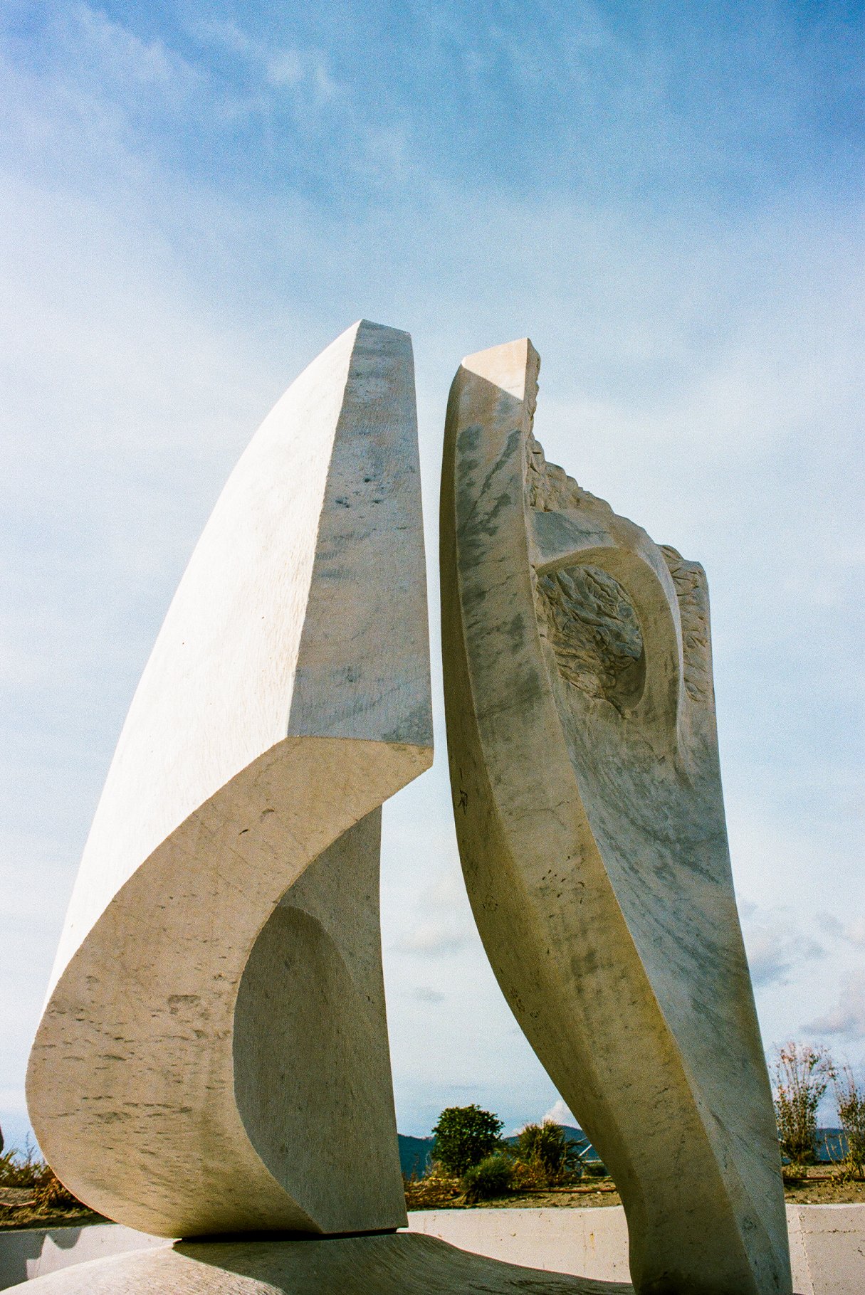 Large abstract sculpture made of concrete with curved and angular shapes, set outdoors against a blue sky with scattered clouds and trees in the background.