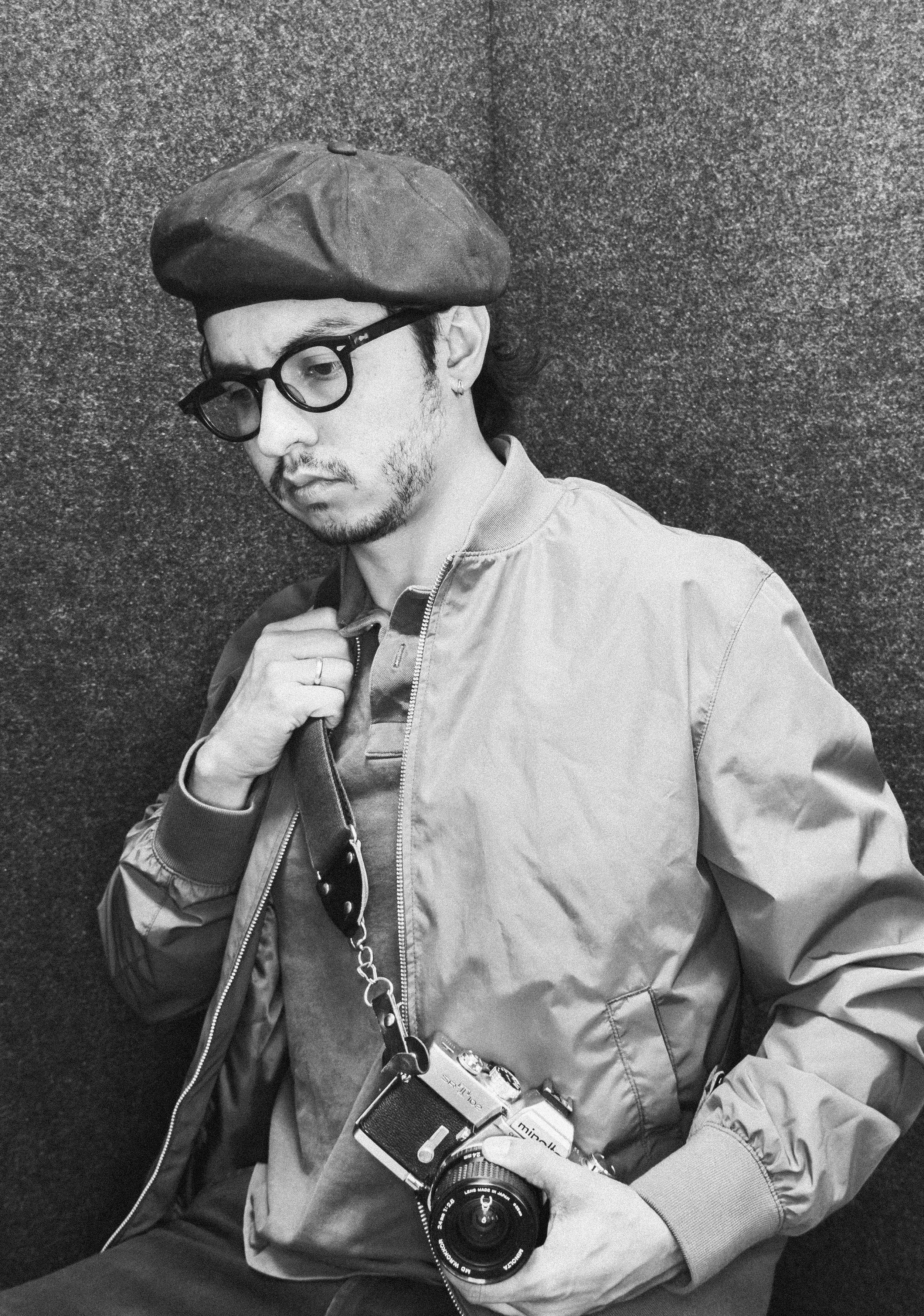 Black and white photo of a young man wearing glasses, a beret, and a jacket, holding a camera with a strap, and standing against a textured wall.