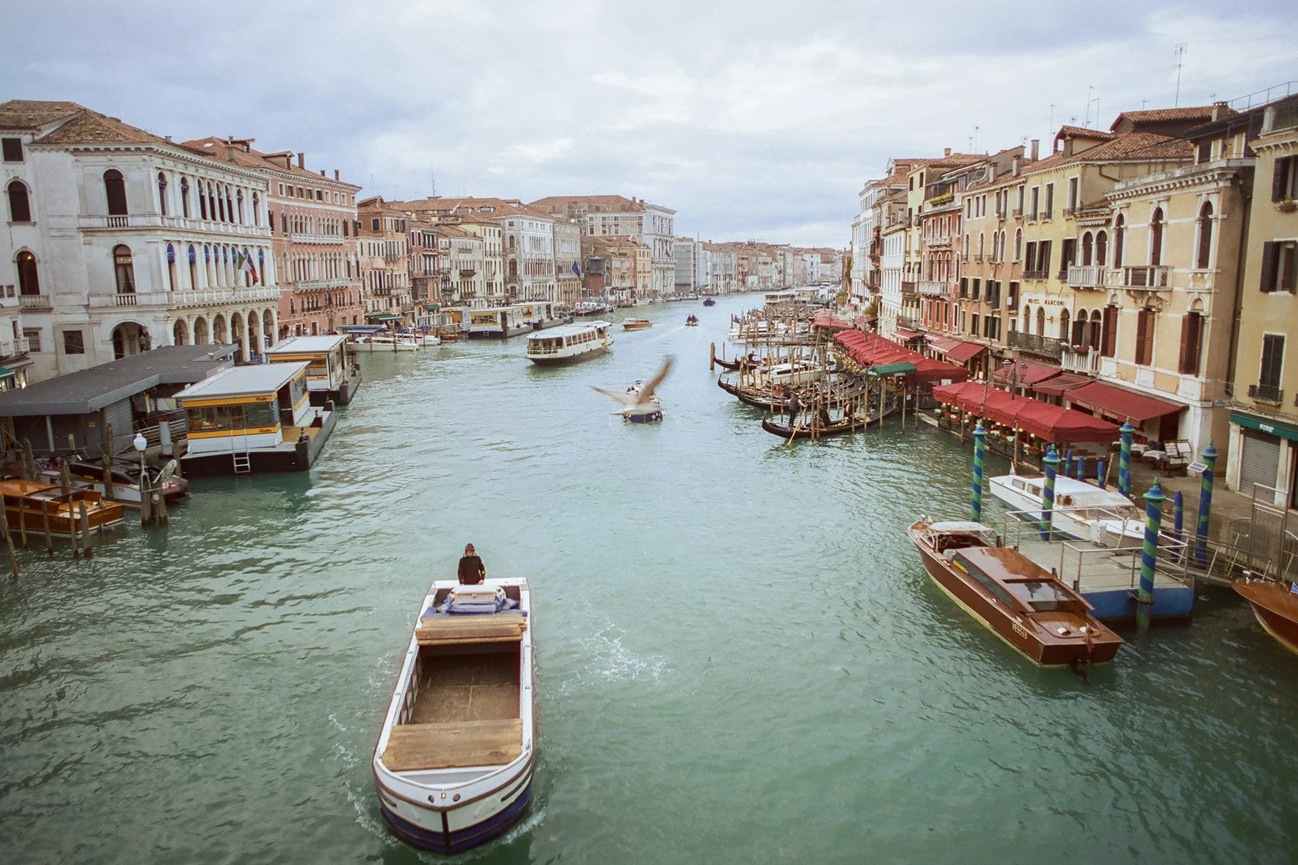 A canal in Venice, Italy with boats, historic buildings with balconies, and outdoor cafes with red umbrellas lining the water's edge.