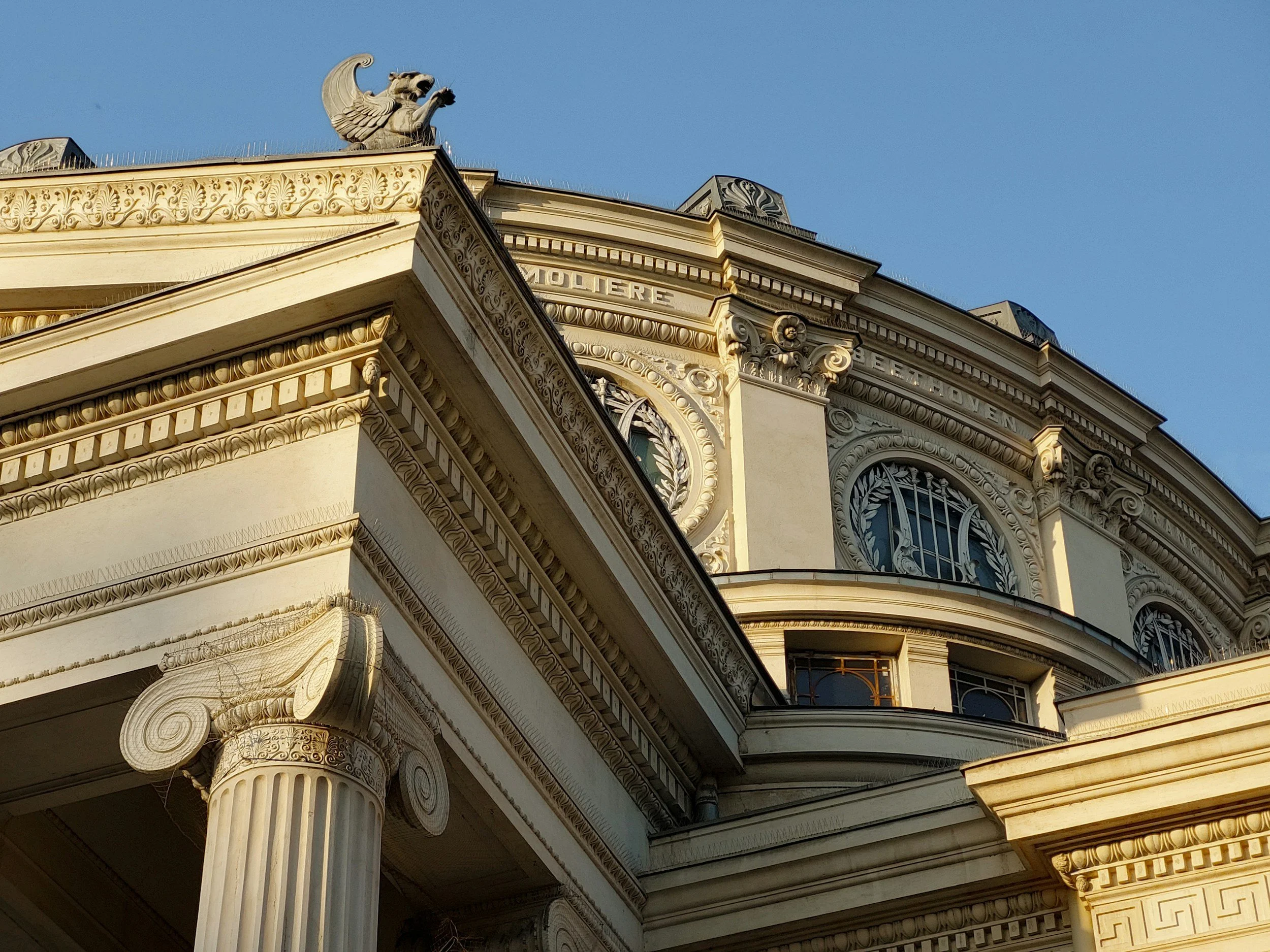 Close-up of ornate neoclassical architectural details on a historic building, including a lion sculpture on the roof, decorative moldings, and Corinthian columns with papyrus plant motifs.