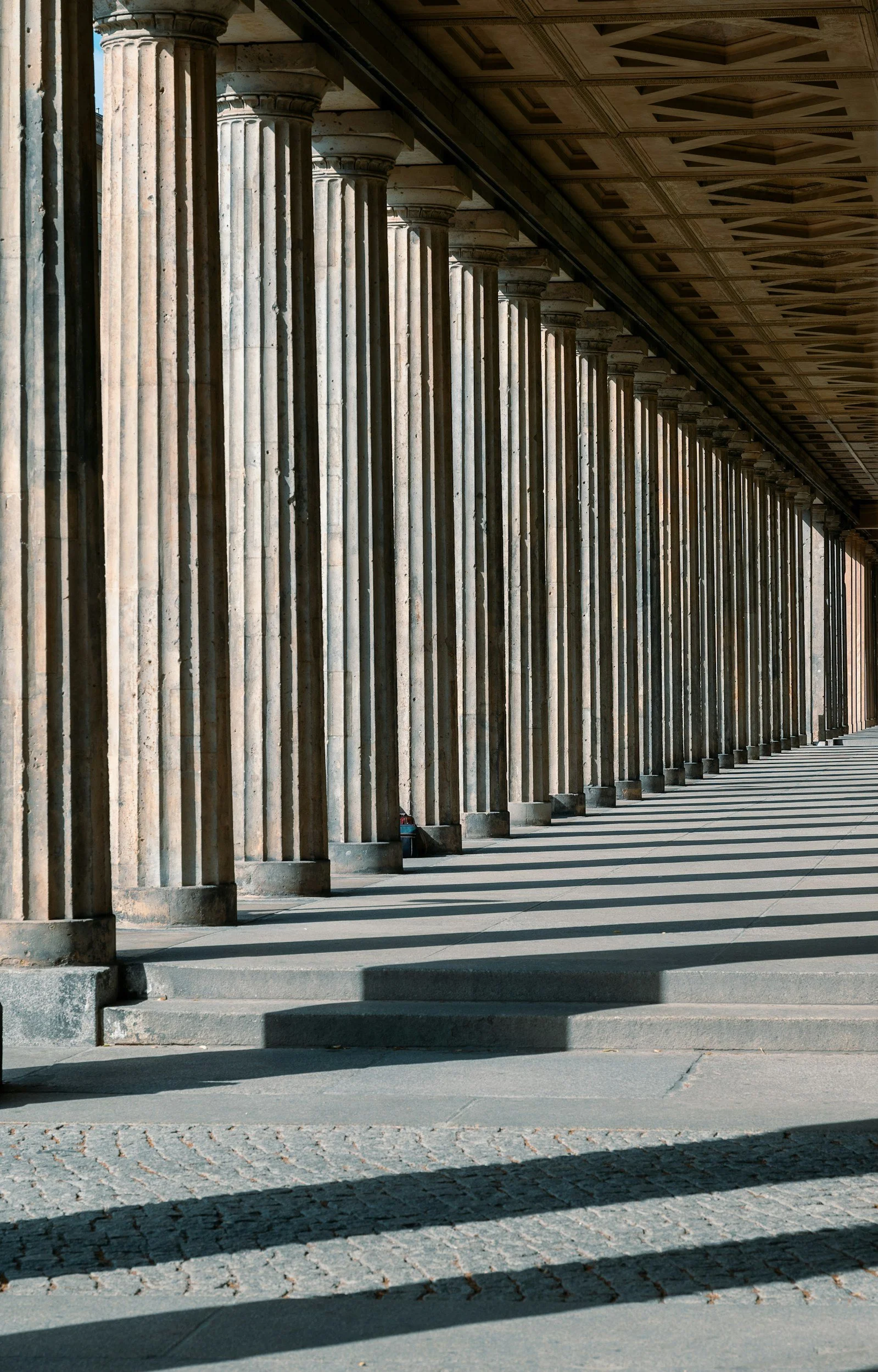 A row of classical stone columns with shadows cast on the ground, under a detailed wooden ceiling in an outdoor corridor.