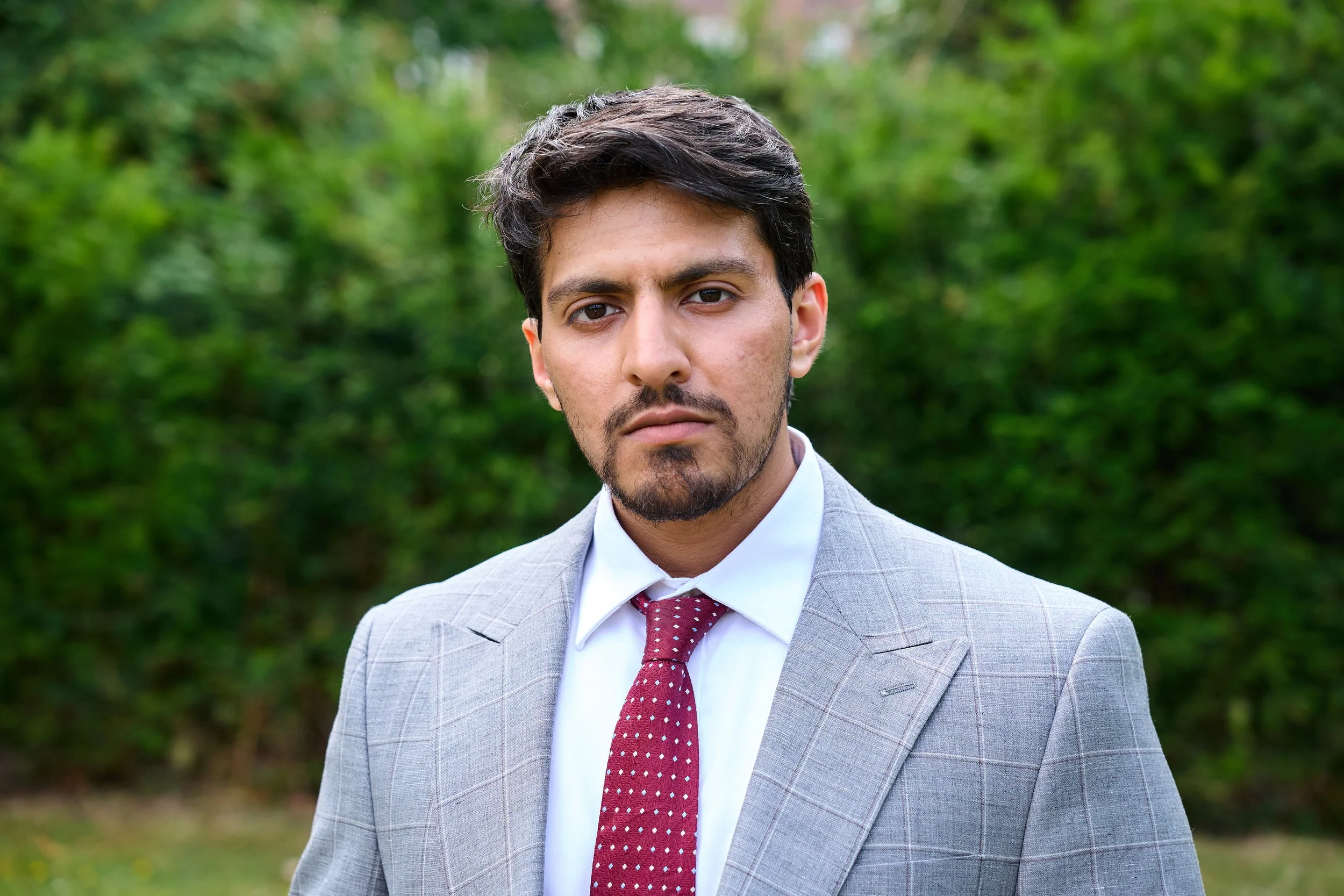 A young man with dark hair and a beard wearing a light gray suit and a red polka dot tie, standing outdoors with a green, blurred background.