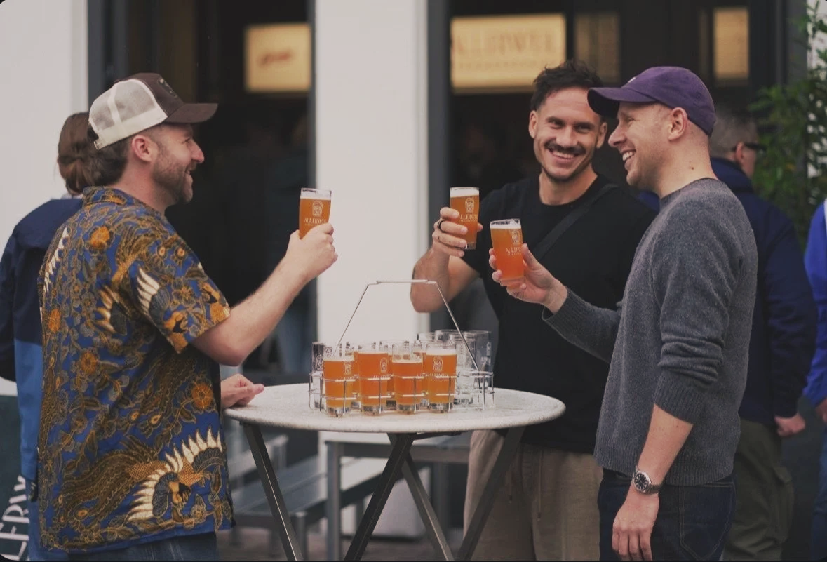 Drei Männer trinken Bier und lachen miteinander, während sie an einem Stehtisch mit mehreren Biergläsern stehen.