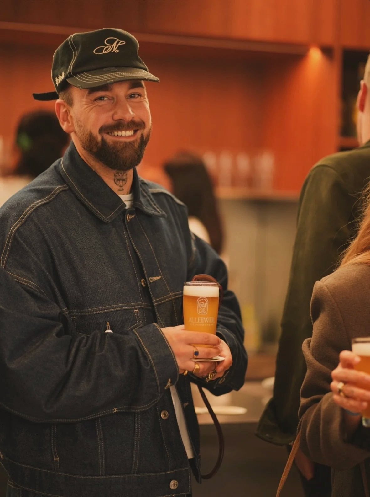 Ein Mann mit Bart und schwarzer Jeans hält ein Glas Bier in einer Bar. Er trägt eine schwarze Mütze mit weißen Stickereien und eine dunkle Jeansjacke. Er lächelt und schaut in die Kamera.