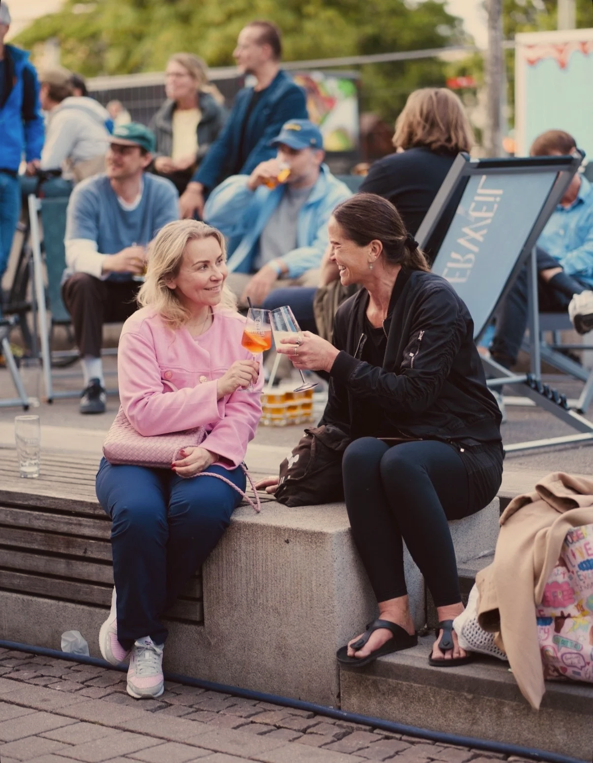 Zwei Frauen sitzen auf einer Bank im Freien, lachen und stoßen mit Gläsern an, umgeben von anderen Menschen bei einem gesellschaftlichen Event.