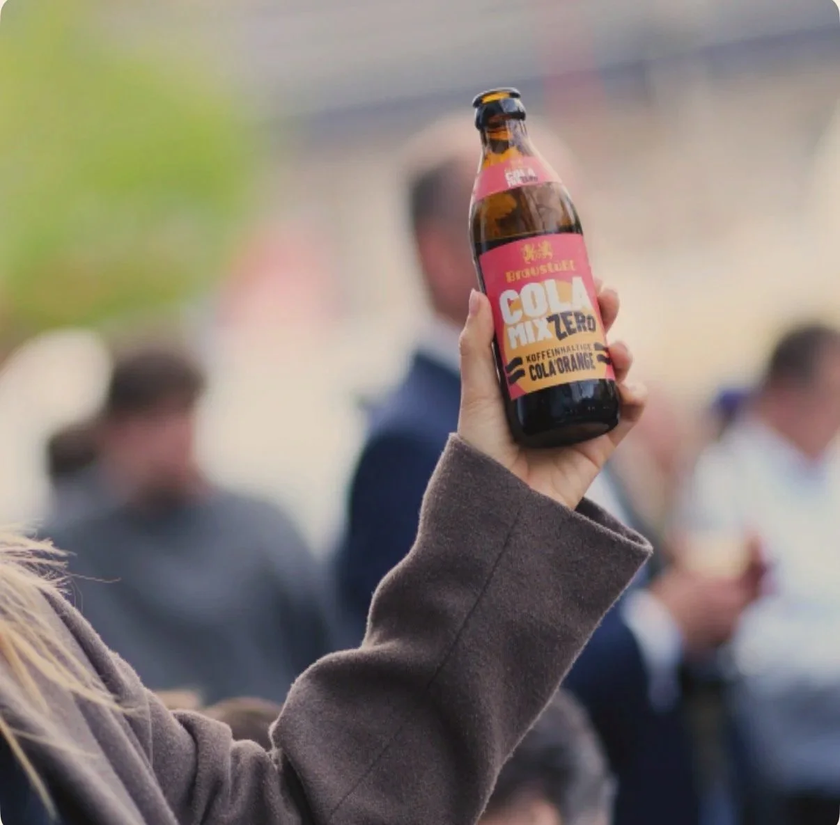 Nahaufnahme einer Hand, die ein Glas Bier mit hellem, orangefarbenem Etikett hochhält, auf dem steht "COLA MIX ZERO" und "KOFFEINFREI" bei einem gesellschaftlichen Treffen im Freien.