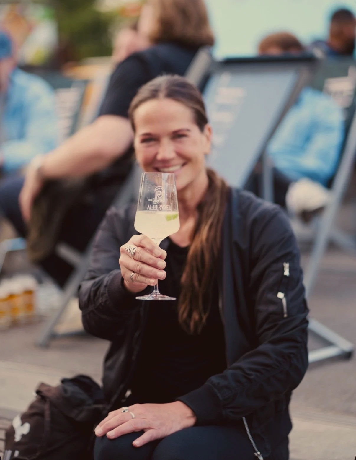 Eine Frau hält ein Weinglas mit weißem Wein bei einer Veranstaltung im Freien. Im Hintergrund sind andere Menschen und Liegestühle. Die Stimmung wirkt entspannt und fröhlich.