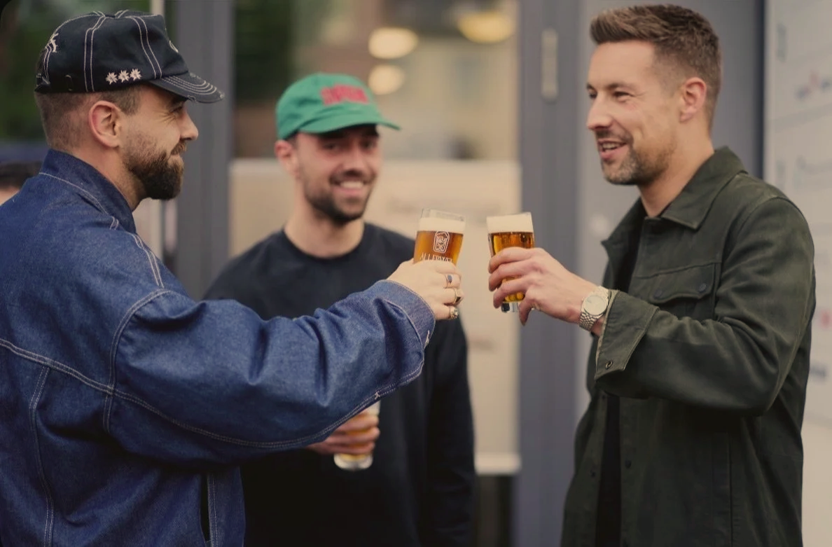 Drei Männer stoßen mit Biergläsern an und lachen in einer geselligen Runde.