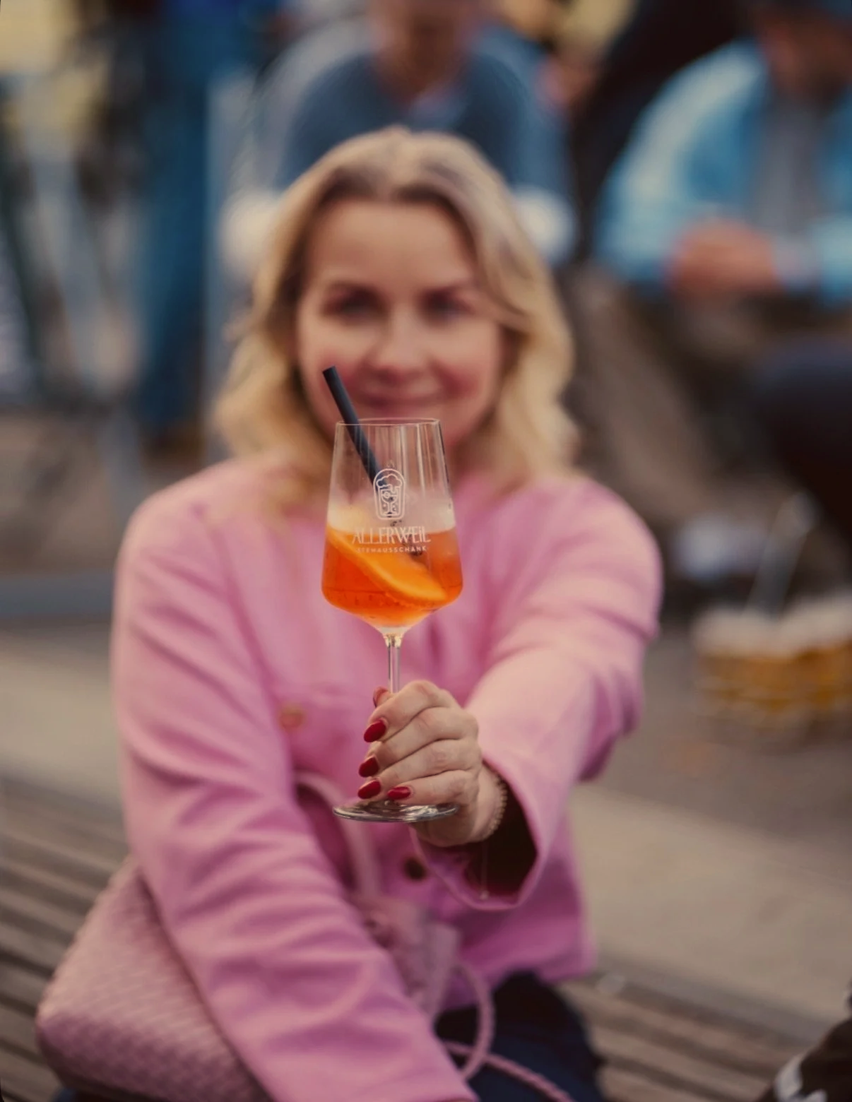 Frau hält ein Aperol Spritz Glas mit einer Orangenscheibe und einem schwarzen Strohhalm, im Hintergrund Menschen in einer Bar.