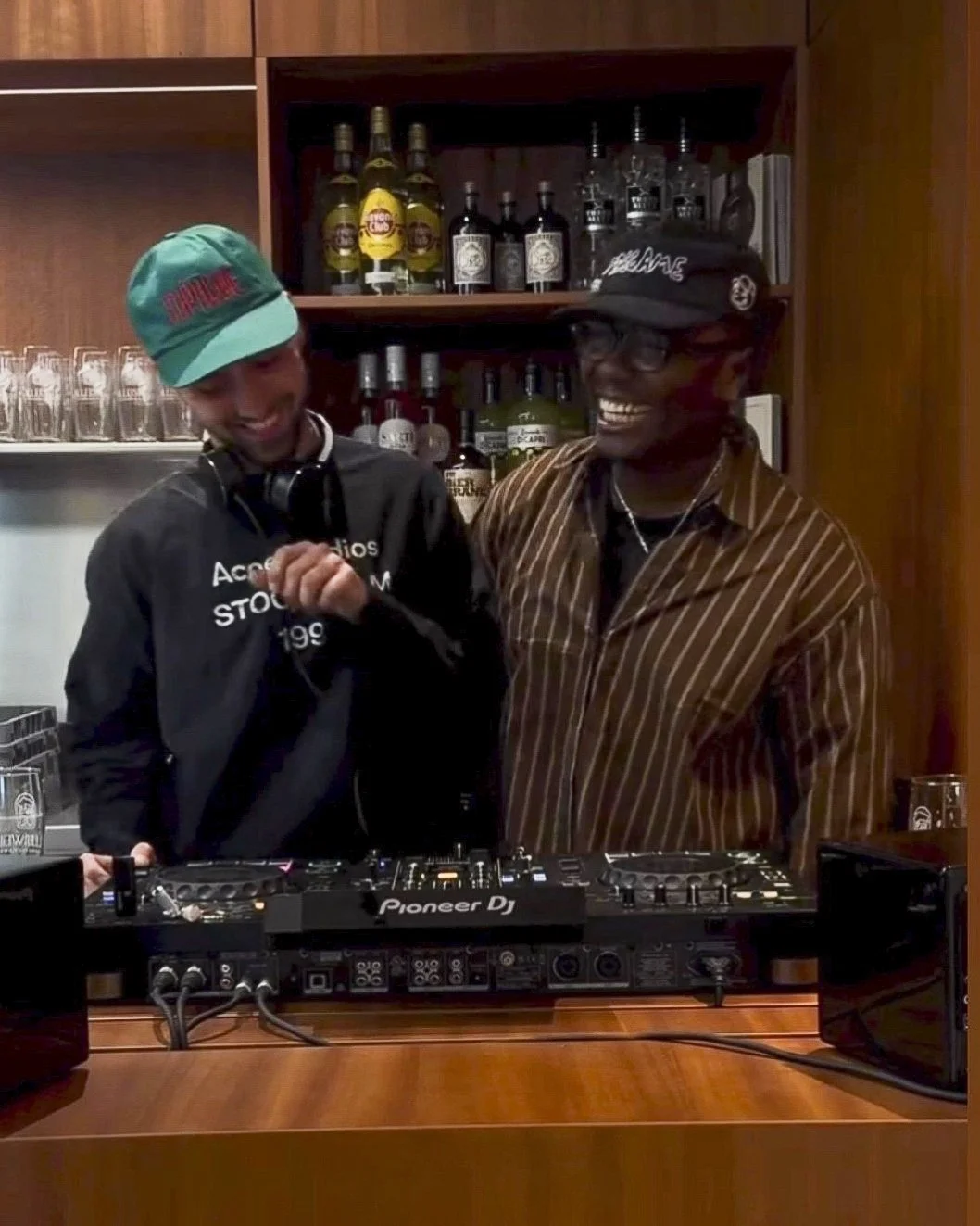 Zwei Männer beim DJen in einer Bar, lächelnd und lachend, mit alkoholischen Getränken im Hintergrund.