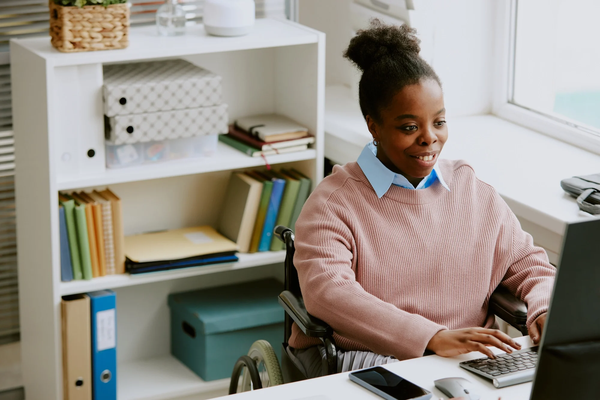 A young woman in a wheelchair working on a laptop at a desk in an office, with bookshelves behind her.