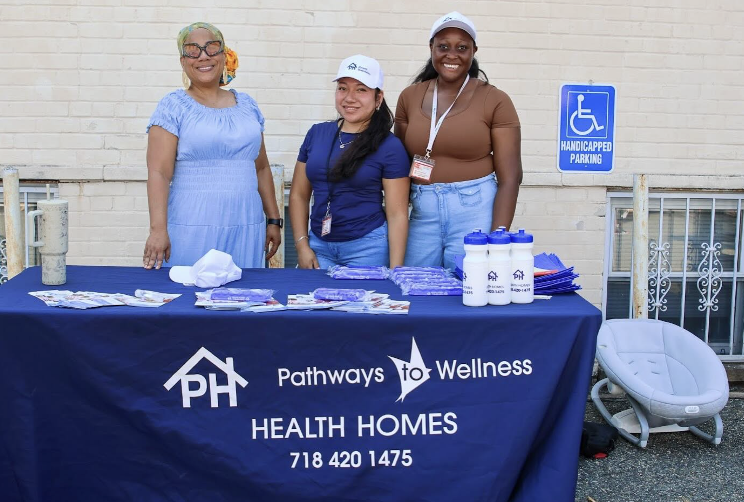 Three women standing behind a table with health brochures and water bottles, promoting pathways to wellness and healthcare services, outdoors against a brick wall, near a handicapped parking sign.