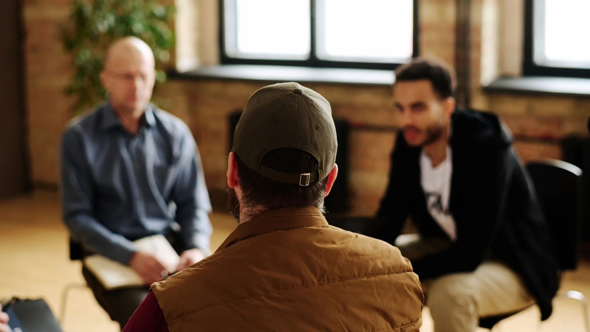 Three men seated in a room with exposed brick walls and large windows, engaged in conversation, with one man in the foreground wearing a green cap and brown jacket, and two men in the background, one blurred with glasses and the other smiling.
