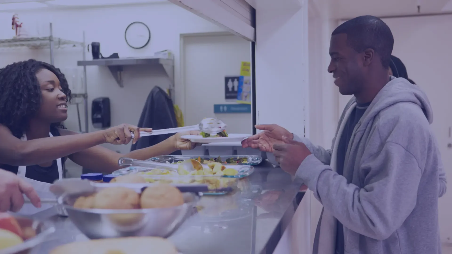 A woman in a hospital kitchen serves food to a man in a gray hoodie. Food trays with salads, bread rolls, and other dishes are on the counter.