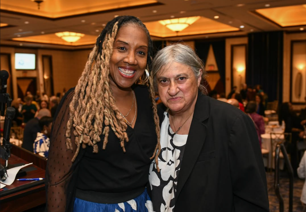 Two women standing together at an indoor event, smiling for the camera, with a crowd in the background.