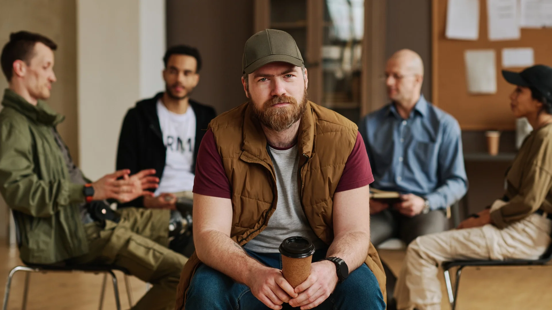 A man with a beard and cap sitting in a room with a group of five diverse people in the background, holding a coffee cup.