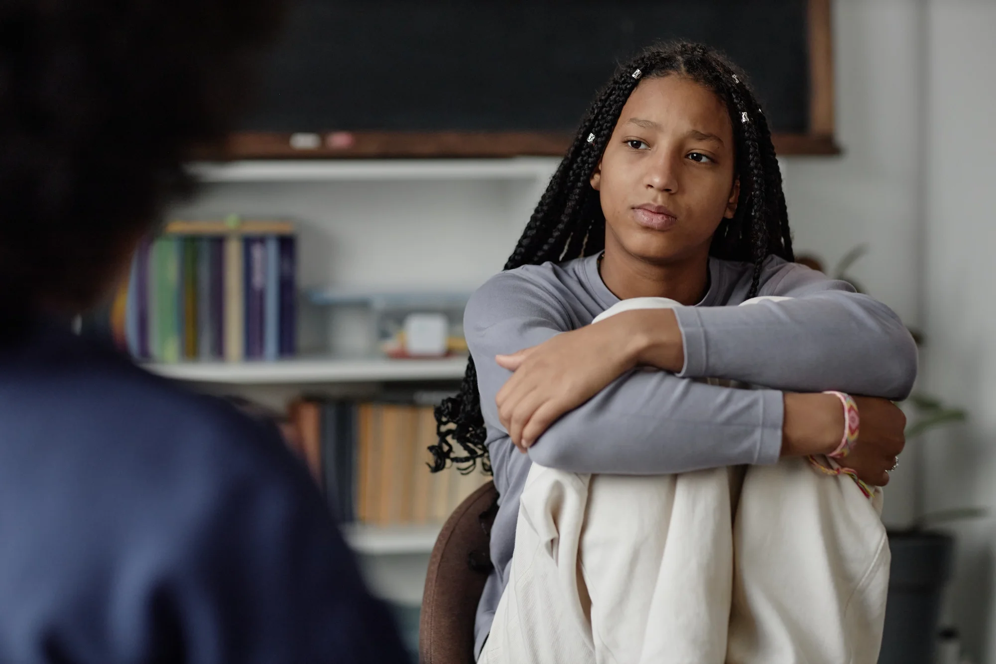 A young girl with braided hair and a bracelet on her wrist sitting in a classroom, looking thoughtfully at someone out of frame, with a blackboard and shelves in the background.