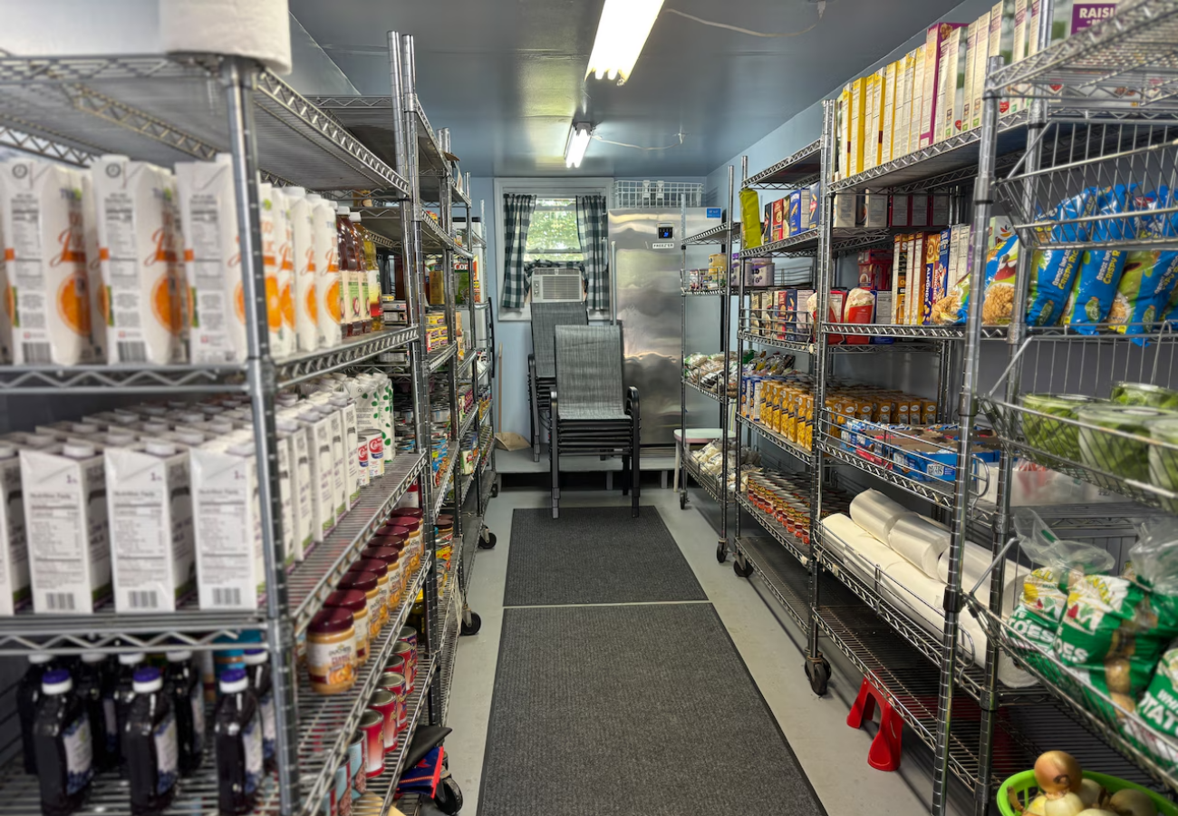 A narrow aisle of a small grocery store with metal shelves on both sides stocked with various food products and snacks. At the end of the aisle, there are stacked chairs near a window with checkered curtains. The floor has black mats, and there are fluorescent ceiling lights.