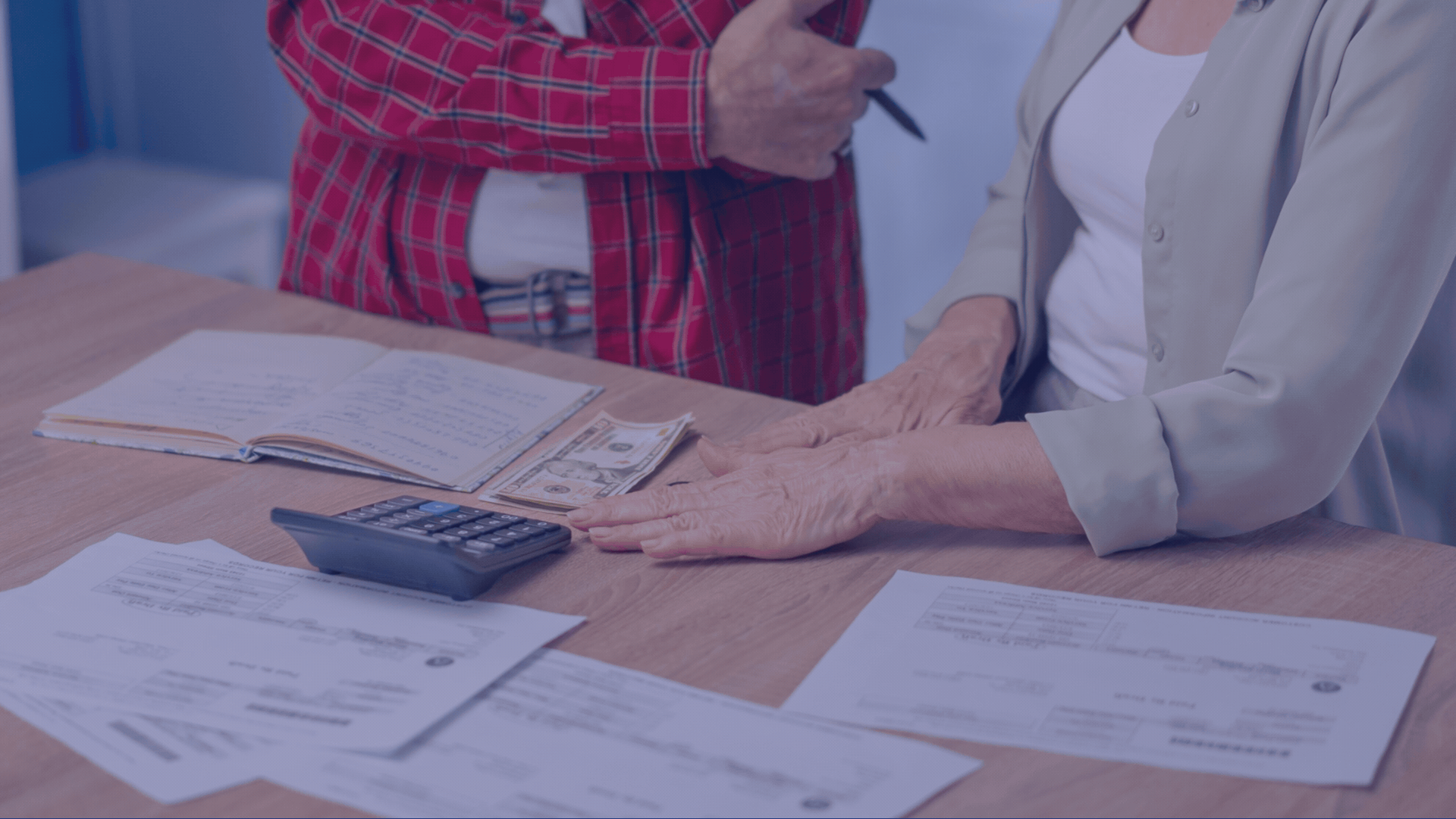 An elderly person and a woman in a beige jacket shaking hands across a table with documents, a calculator, and cash on it.