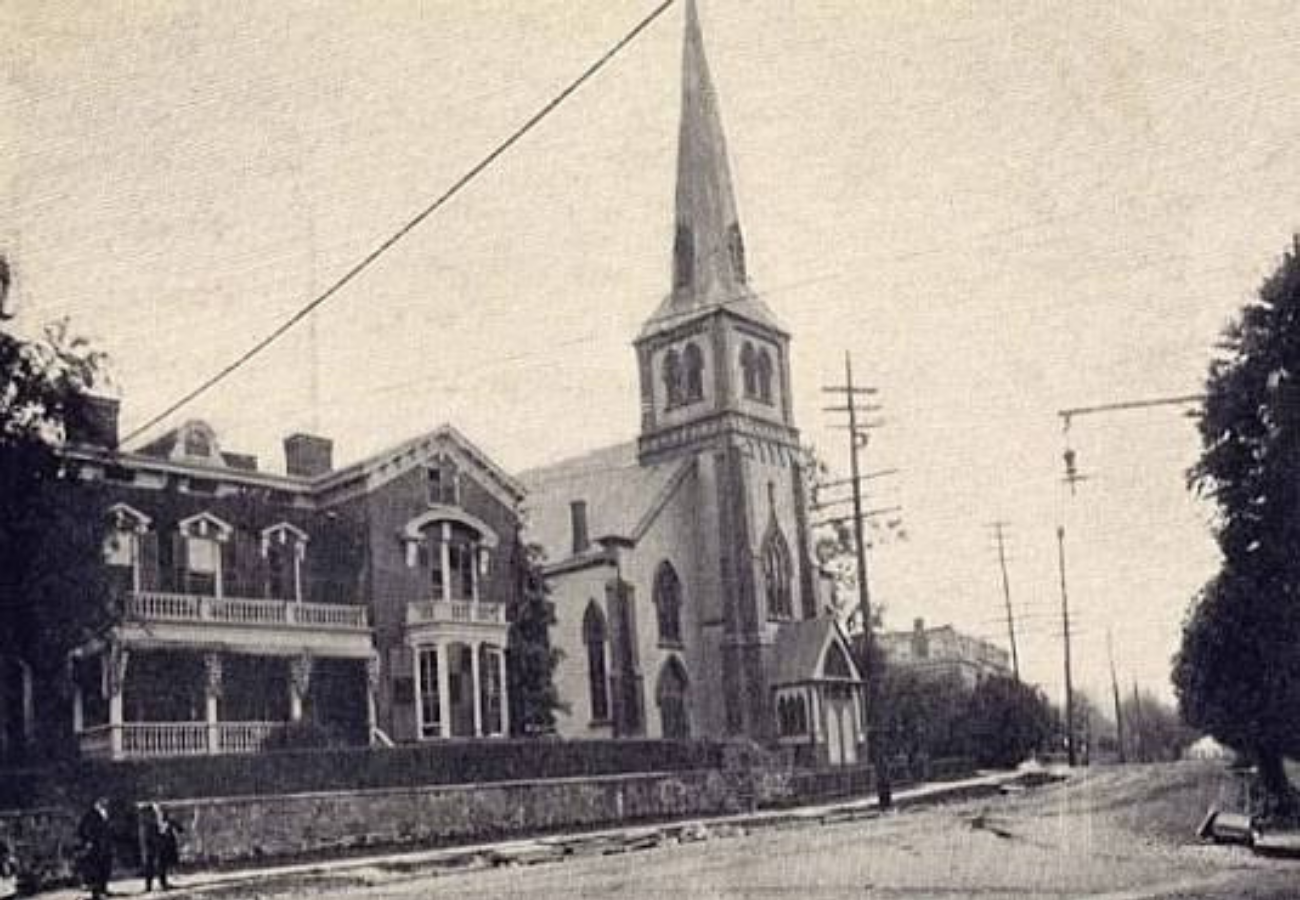 Black and white photo of a church with a tall steeple next to a large house with a porch, utility poles line the street, and a few people walking on the sidewalk.