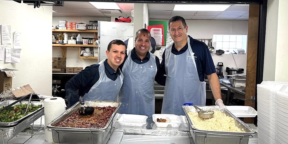 Three men standing behind a kitchen counter with food trays, smiling at the camera, wearing aprons.