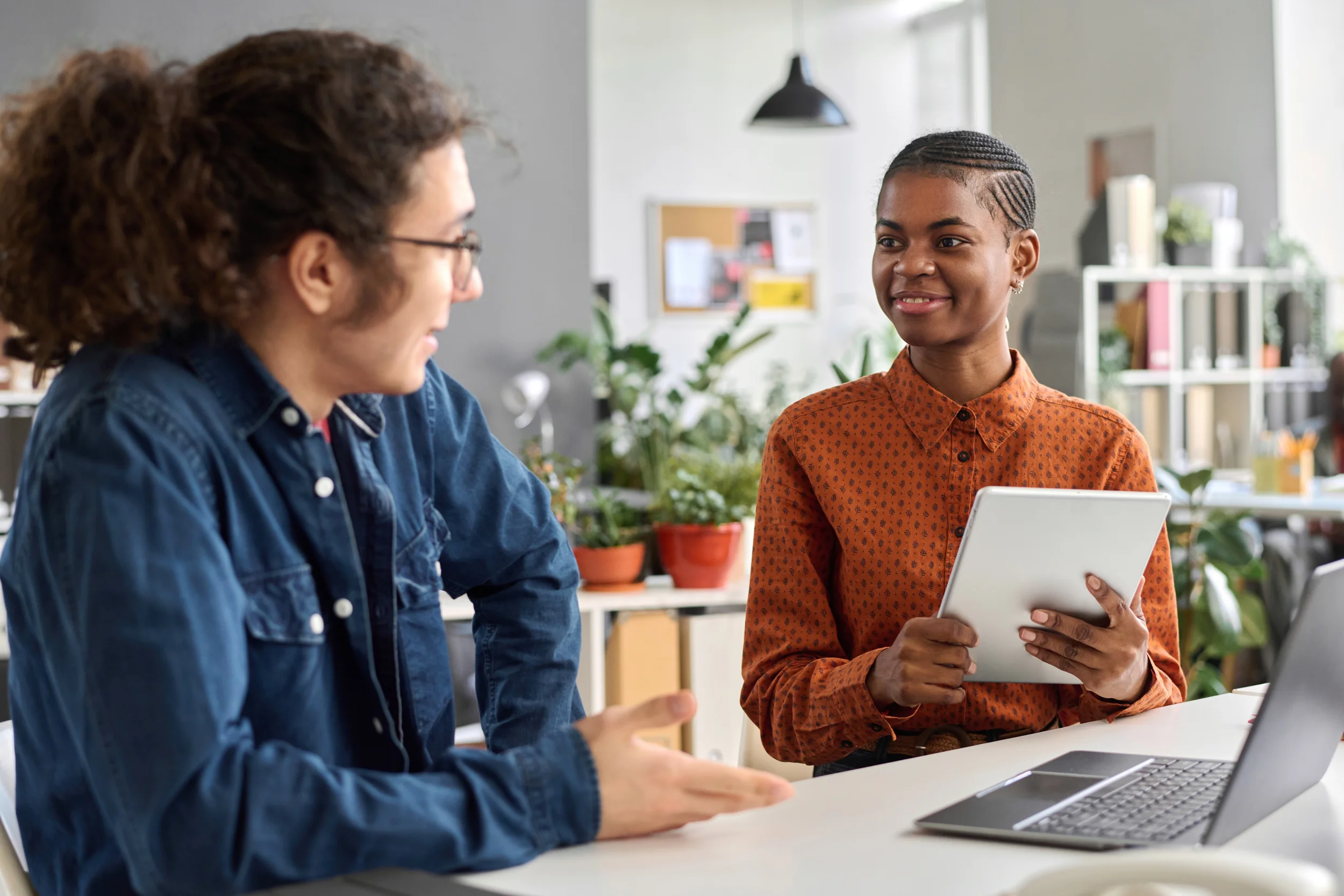 Two women having a conversation in an office. One woman is wearing glasses and a blue denim shirt, while the other woman is holding a tablet and wearing an orange patterned shirt with braids in her hair. The workspace is bright with plants and shelves in the background.
