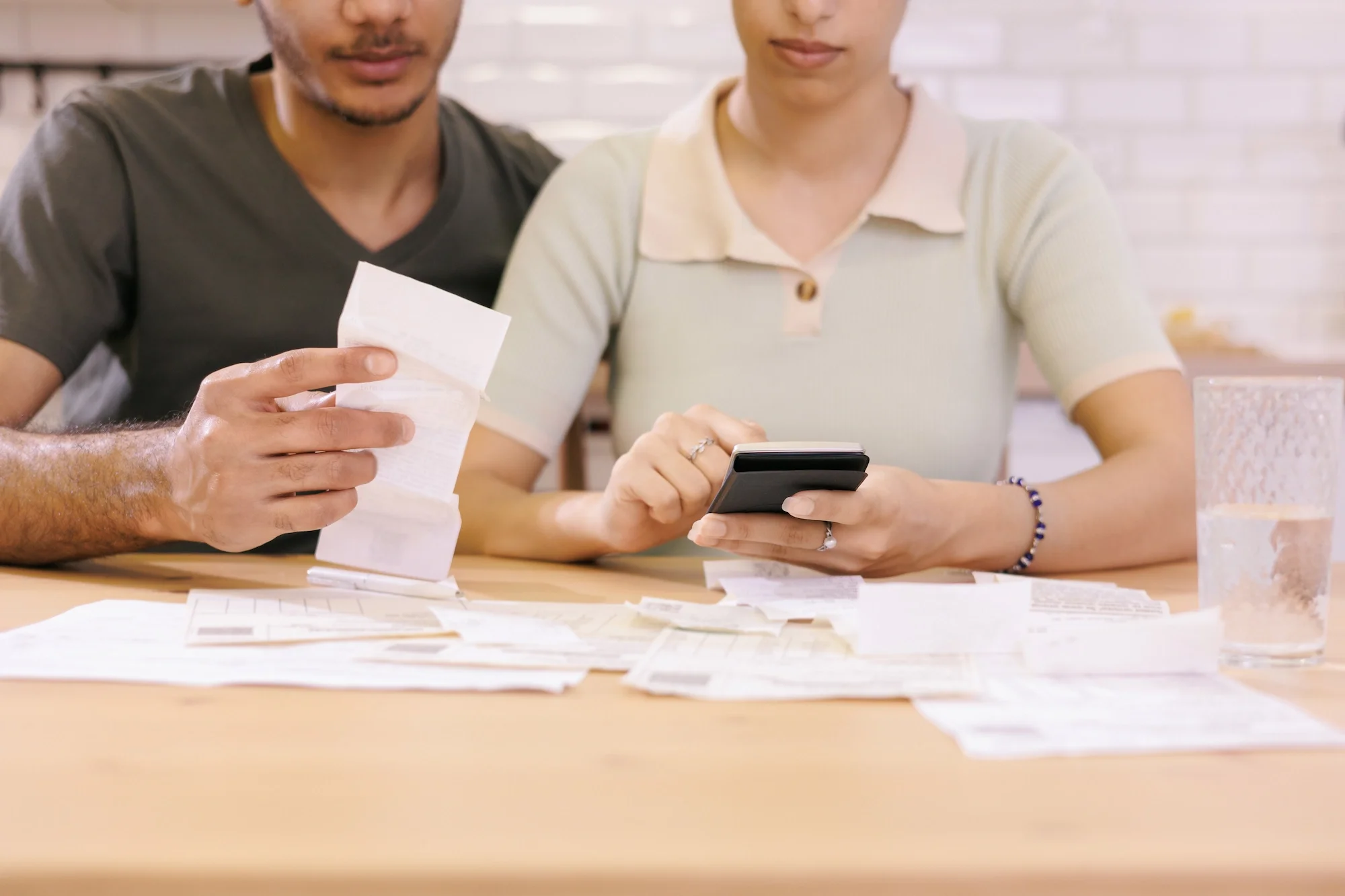 Two people sitting at a table looking at receipts and a smartphone, with scattered receipts and papers on the table.