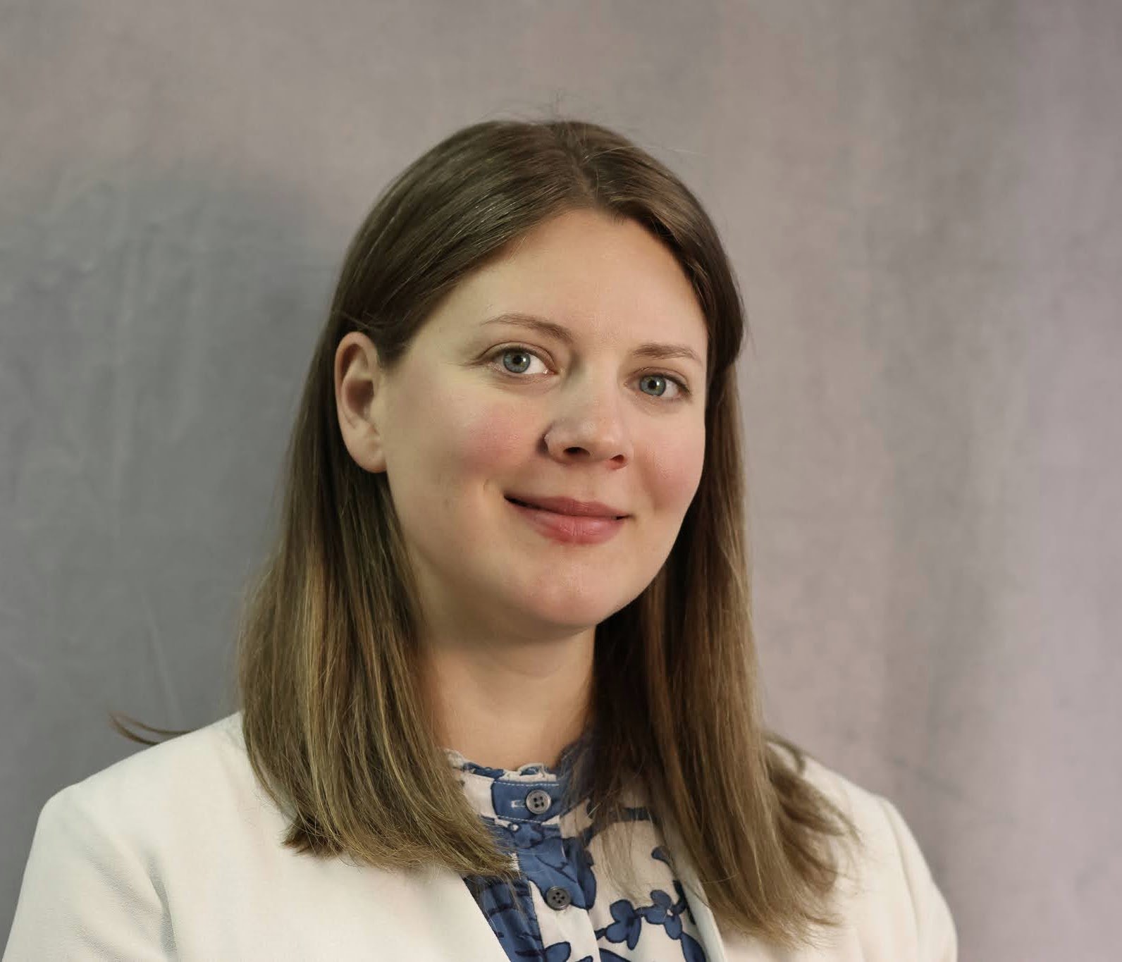 A portrait of a young woman with straight, light brown hair, fair skin, and blue eyes, smiling softly, wearing a cream-colored blazer over a blue and white patterned top, standing against a plain gray background.