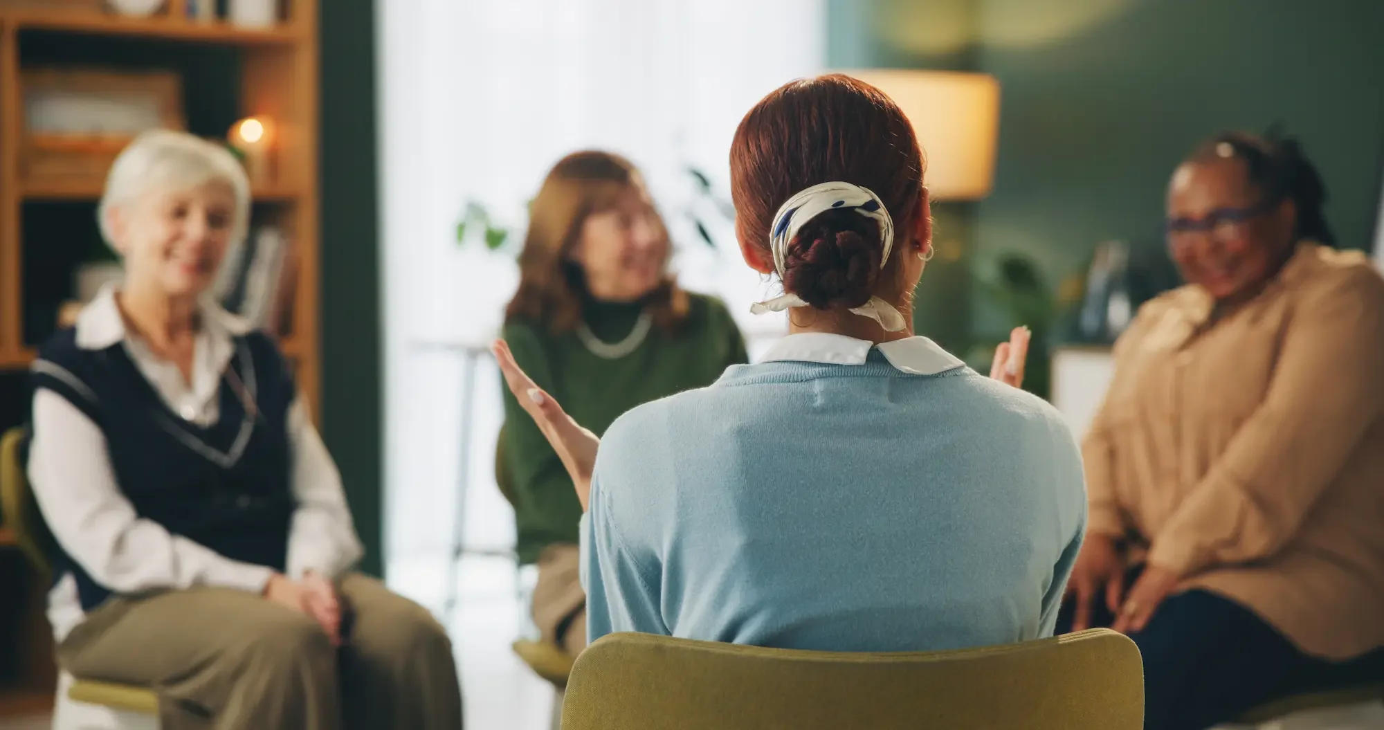 A woman with red hair and a white headscarf sitting in a group session, gesturing with her hands. Three other women are seated around her, listening attentively in a cozy, well-lit room.