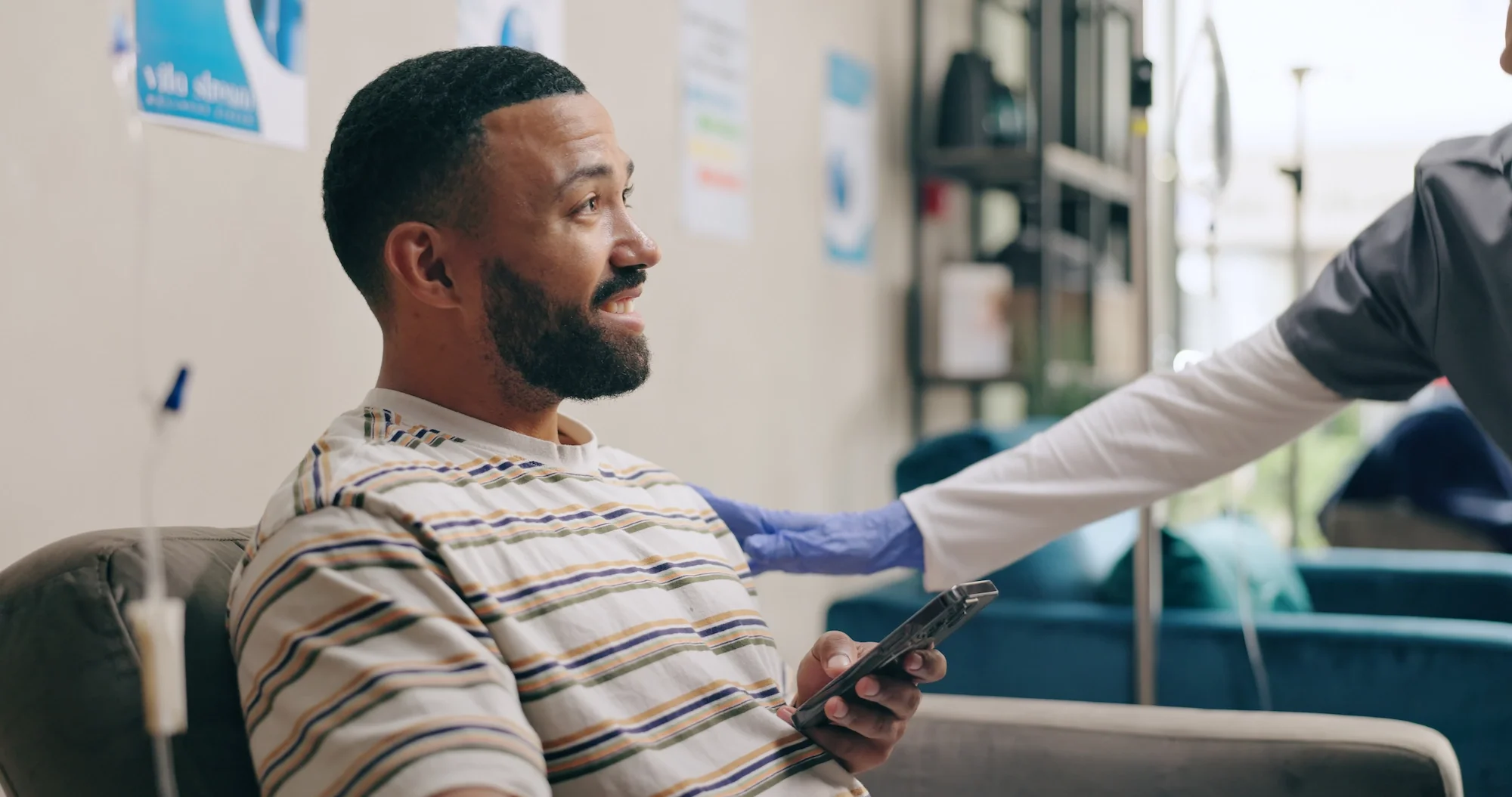 A man with a beard and striped shirt sitting on a chair in a medical facility, holding a smartphone, with a healthcare worker in scrubs placing their hand on his shoulder. There are medical posters and equipment in the background.