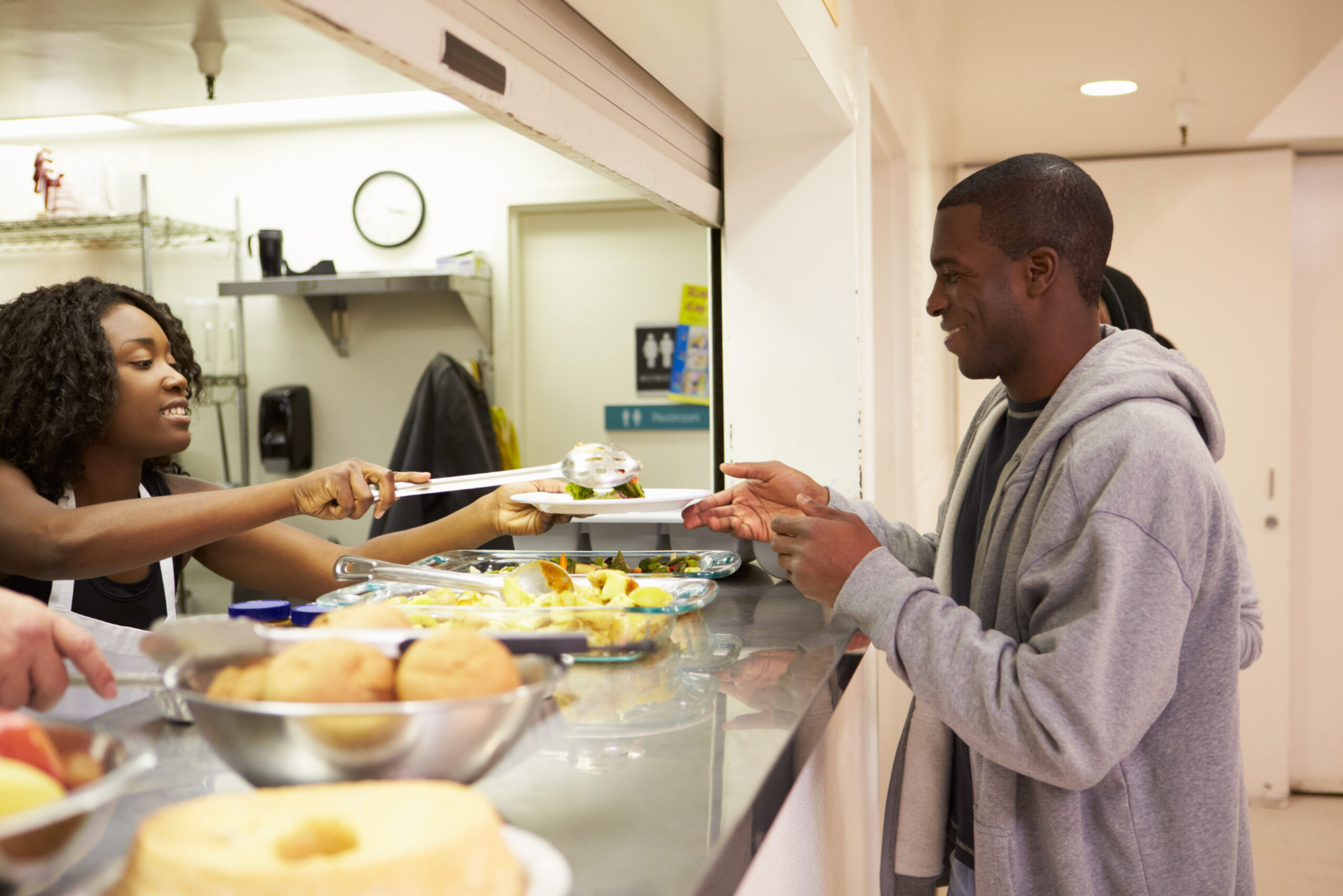 A man in a gray hoodie receives food on a plate from a woman working at a cafeteria serving counter. The counter has various food items, and the woman is using tongs to hand over a plate.