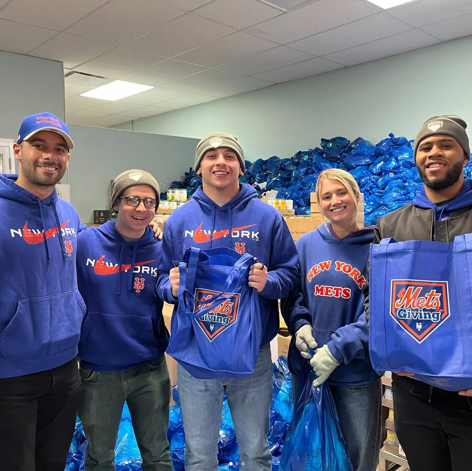 A group of five volunteers wearing blue hoodies and beanies, standing in front of stacks of blue bags and boxes filled with donated goods, participating in a charity event for New York Mets