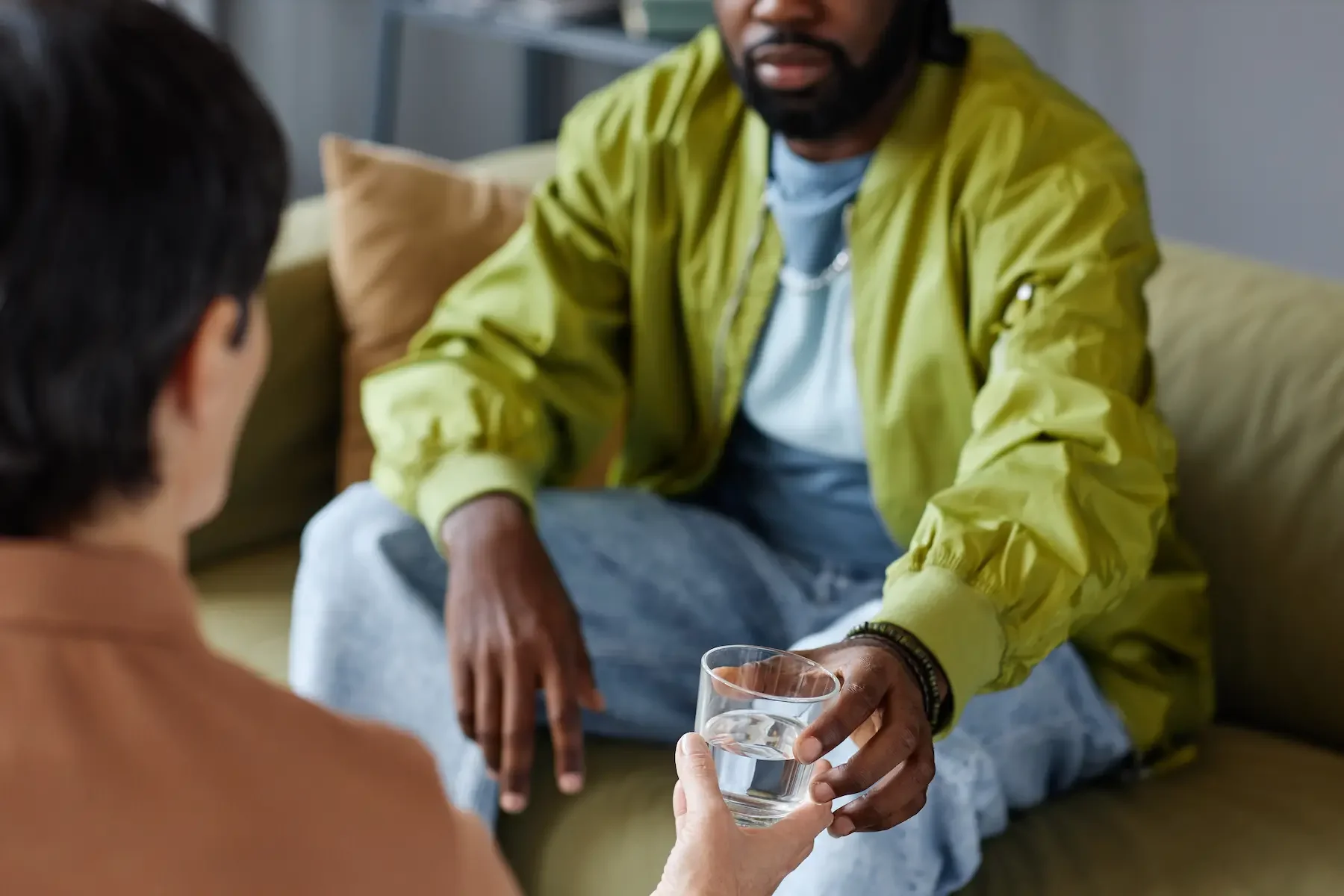 Two people are sitting on the floor, engaging in a conversation. One person is handing a glass of water to the other.
