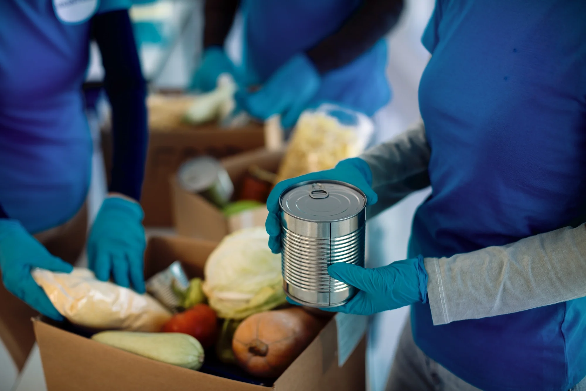 People wearing blue gloves packing canned food, vegetables, and fruits into a cardboard box