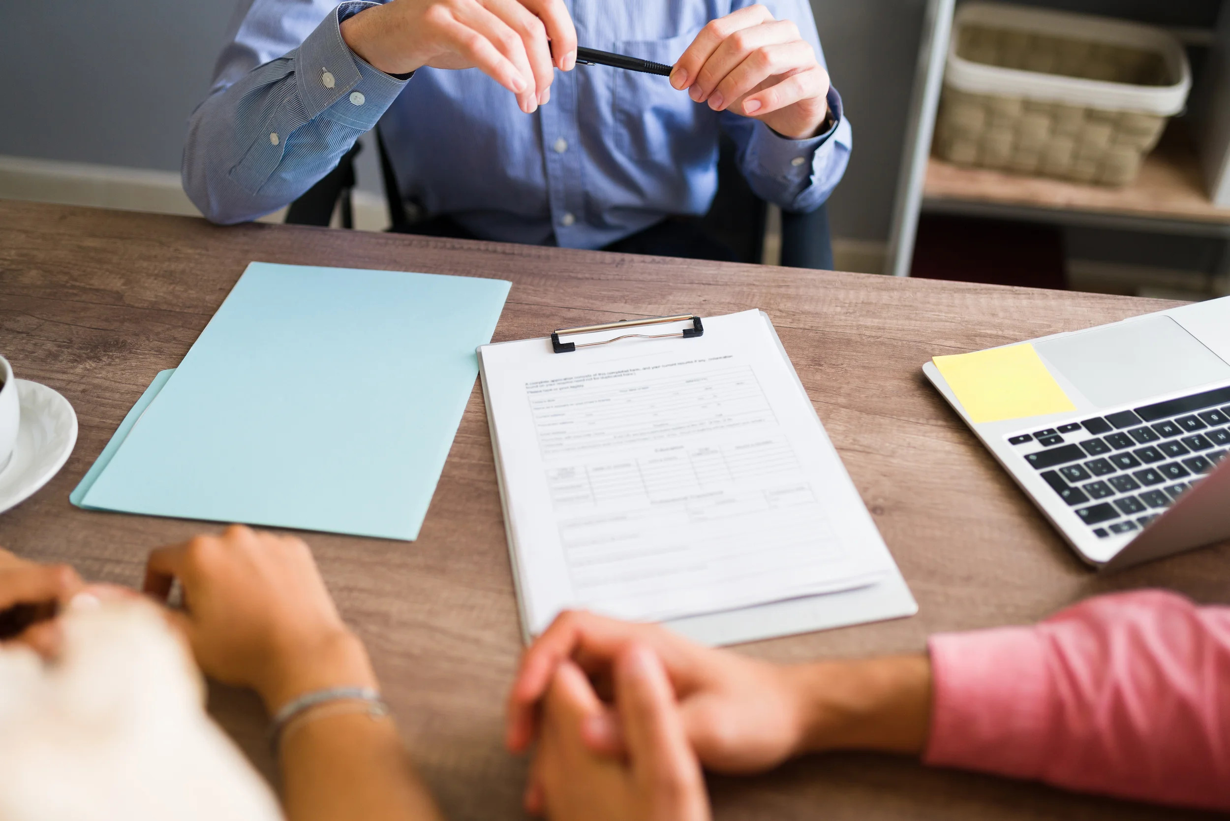 Person in a blue shirt sitting at a wooden desk, holding a pen. On the desk are paperwork on a clipboard, a laptop, a light blue folder, and a white dish. Another person in a pink sleeve is visible in the foreground.