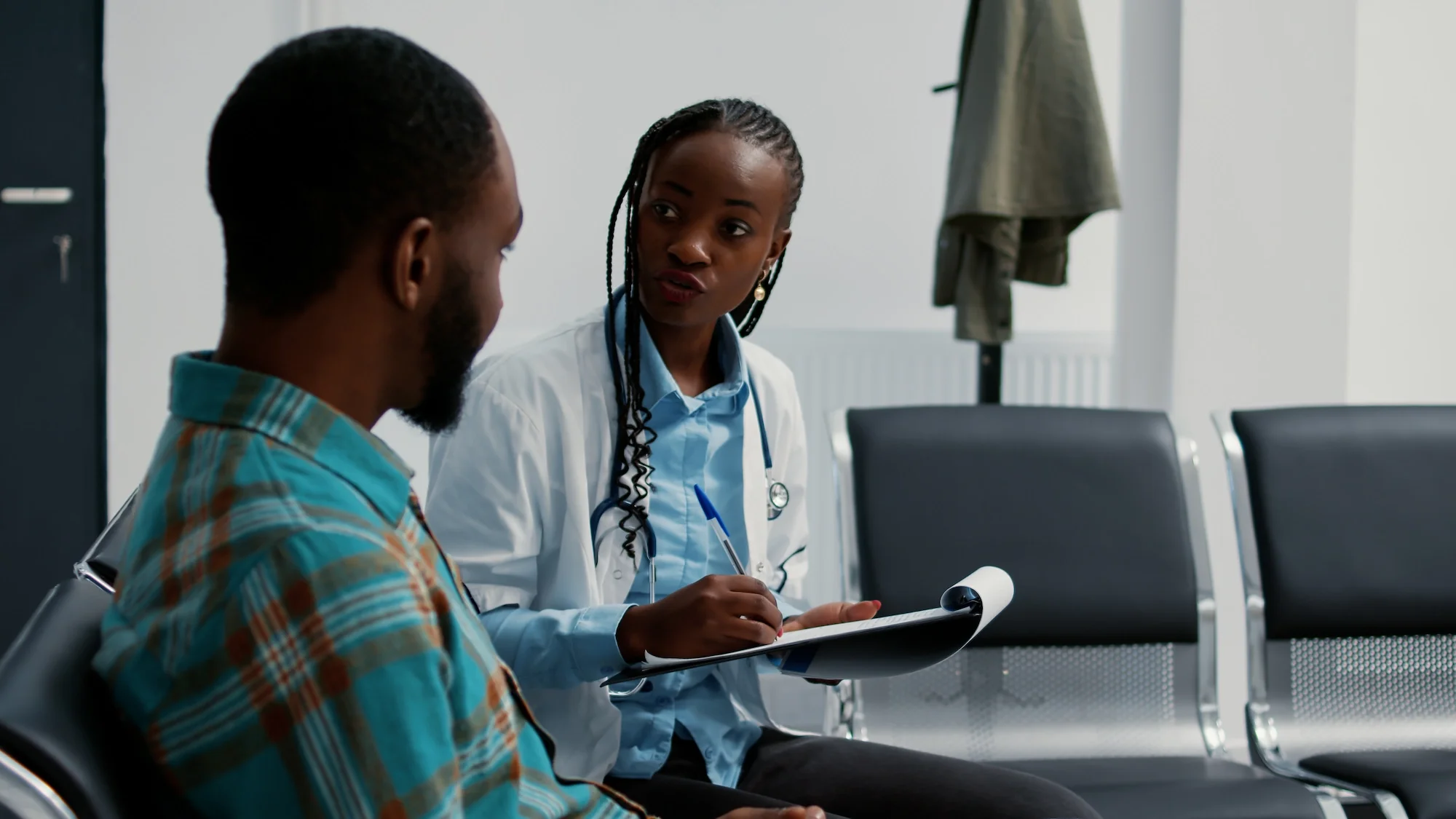 A female doctor in a white lab coat with a stethoscope around her neck talking to a male patient in a waiting room. The doctor is holding a notepad and pen, and there are chairs and a coat rack in the background.