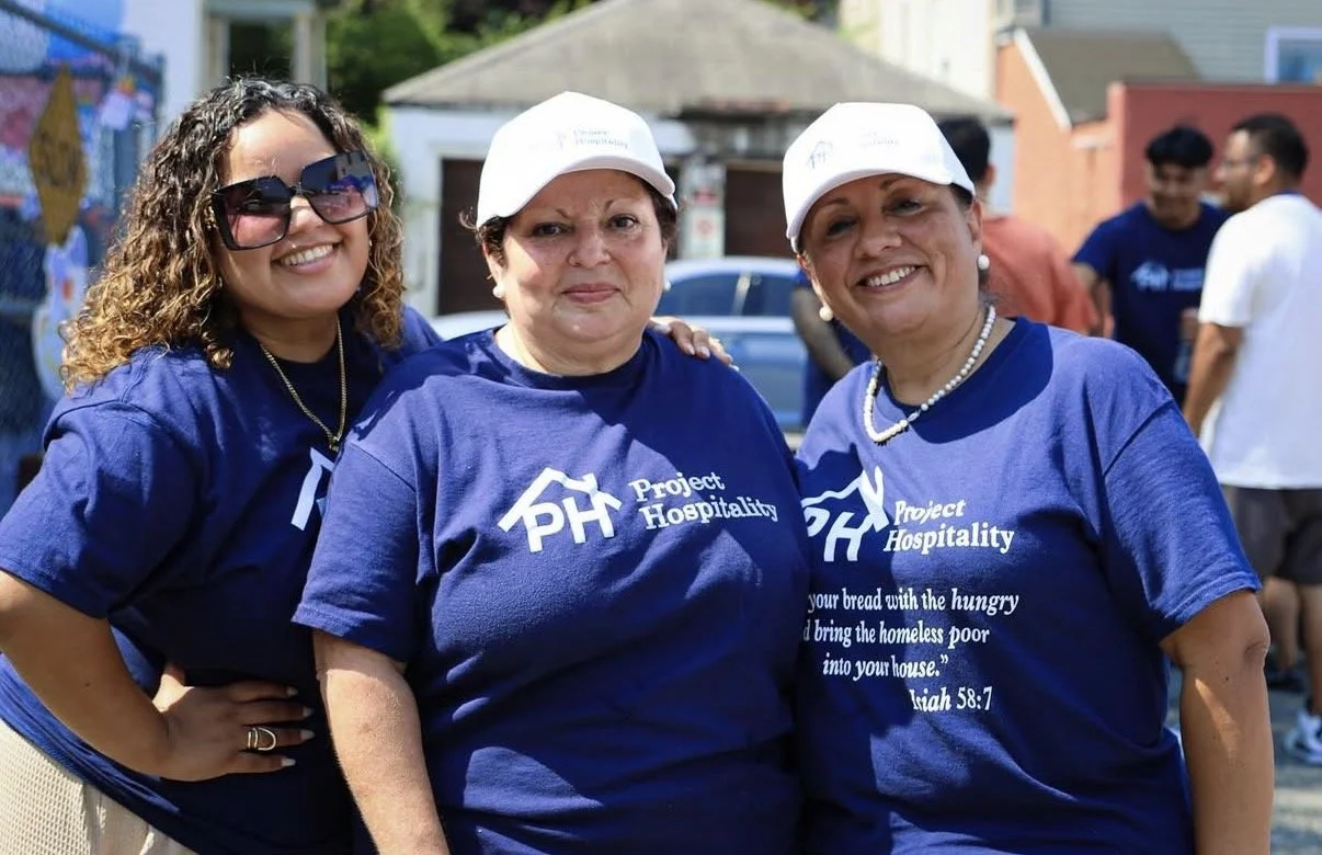 Three women wearing matching blue t-shirts with 'Project Hospitality' logo, standing outdoors on a sunny day, smiling at the camera.