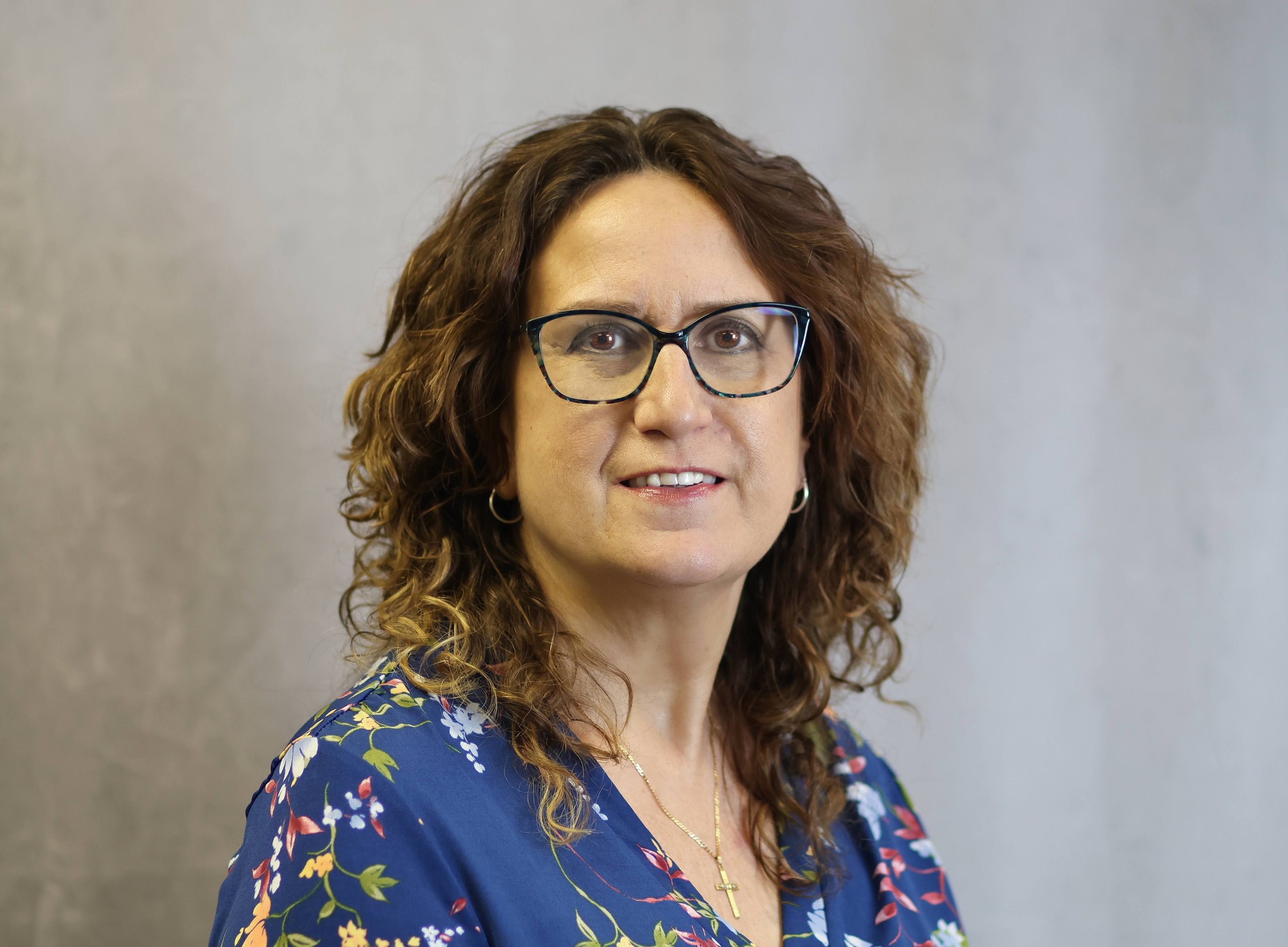 A woman with curly brown hair wearing glasses and a blue floral dress, standing against a plain background.