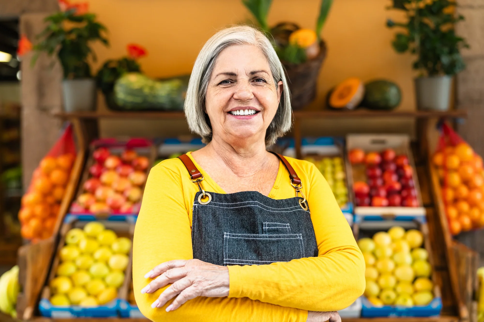 Smiling elderly woman with short gray hair standing in front of a display of apples and pumpkins at a produce store, wearing a yellow long-sleeve shirt and a black apron with brass buckles.