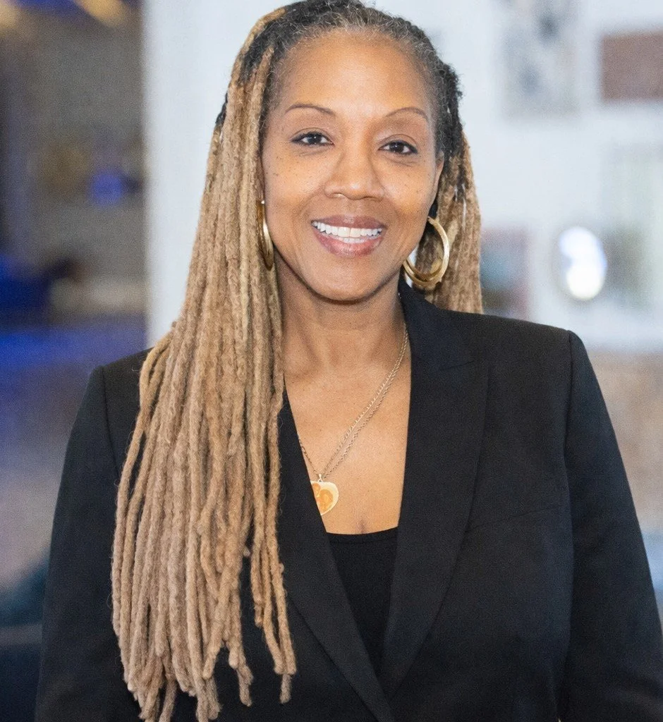 A woman with long blonde dreadlocks, wearing a black blazer, gold hoop earrings, and a necklace, smiling at the camera.