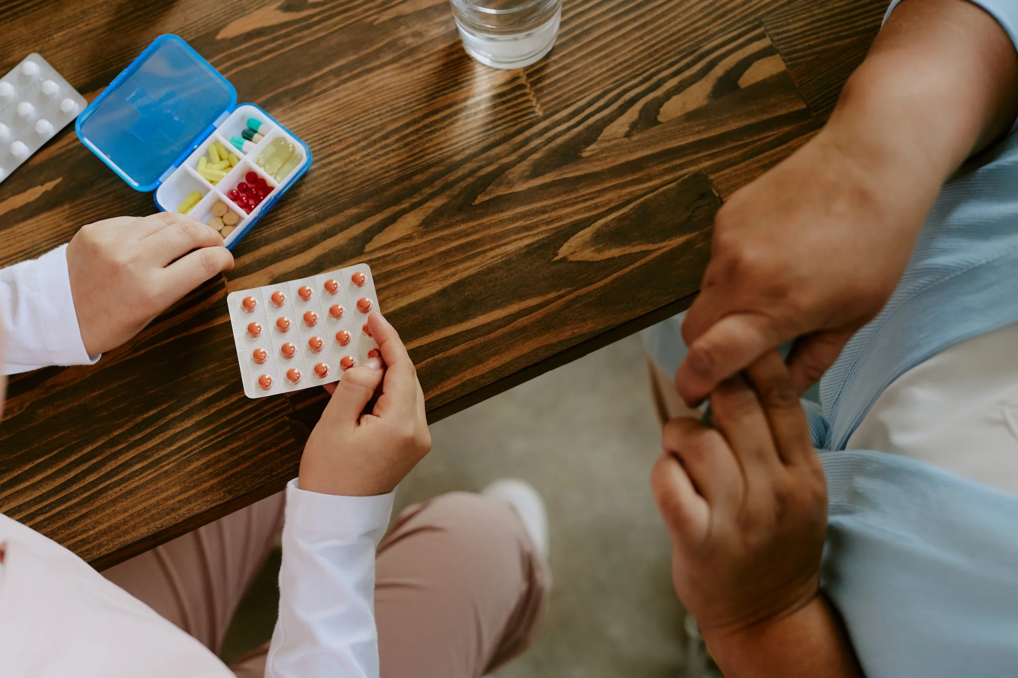 Child and elderly person hold hands across a wooden table, with medication and a glass of water nearby.